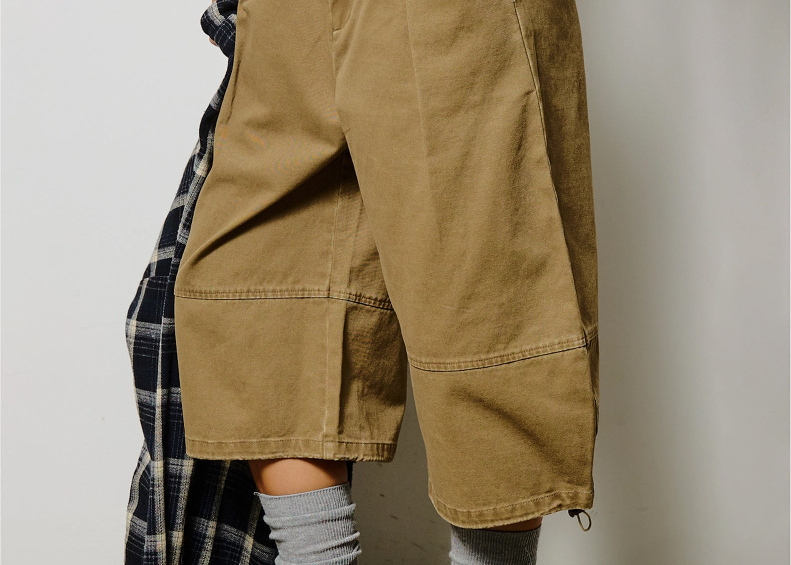 A person wearing oversized cropped utility pants in a warm khaki-tan color, styled with gray socks, fuzzy tan slip-on shoes, and a plaid shirt draped over one leg against a plain light gray wall.
