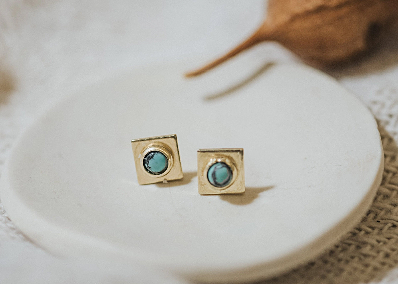 A pair of gold square stud earrings with round turquoise-blue stones, displayed on a small white ceramic dish with a blurred natural background.