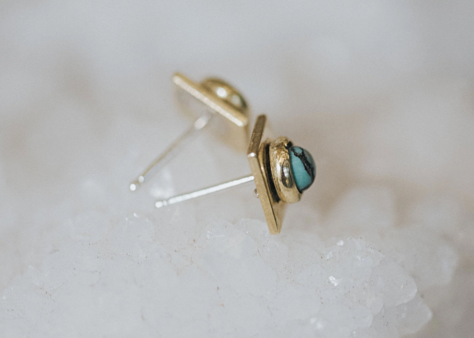 A pair of gold geometric stud earrings with a small round turquoise stone, resting on a white crystal surface.