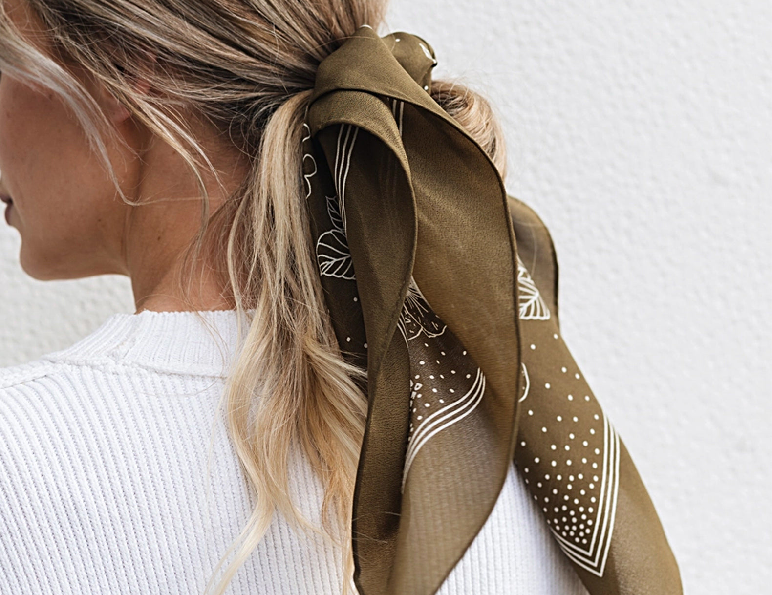 A woman shown from the back wearing a brown silk scarf tied around her hair, with a white ribbed sweater against a textured white wall.