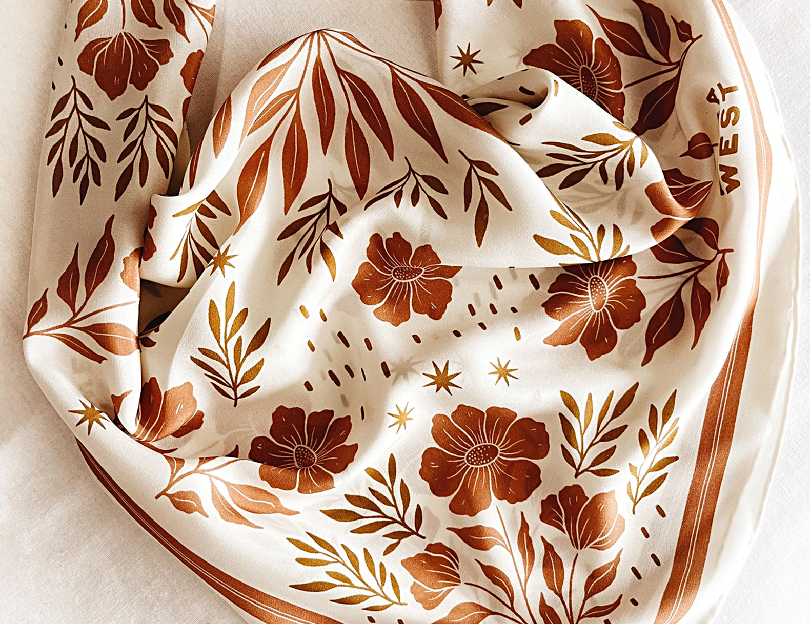 A square cream scarf with a brown floral and botanical pattern, folded and draped on a white textured fabric background. The brand name 'WEST' is printed in brown on one corner.