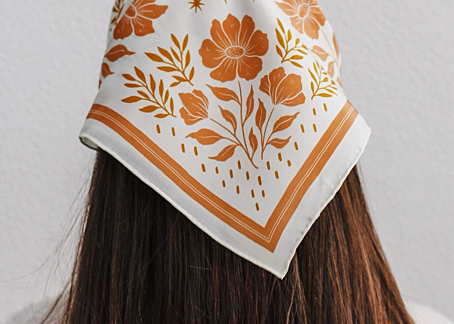 A woman viewed from behind wearing a square white scarf with an orange floral and botanical print, styled over her dark brown hair against a light gray wall.