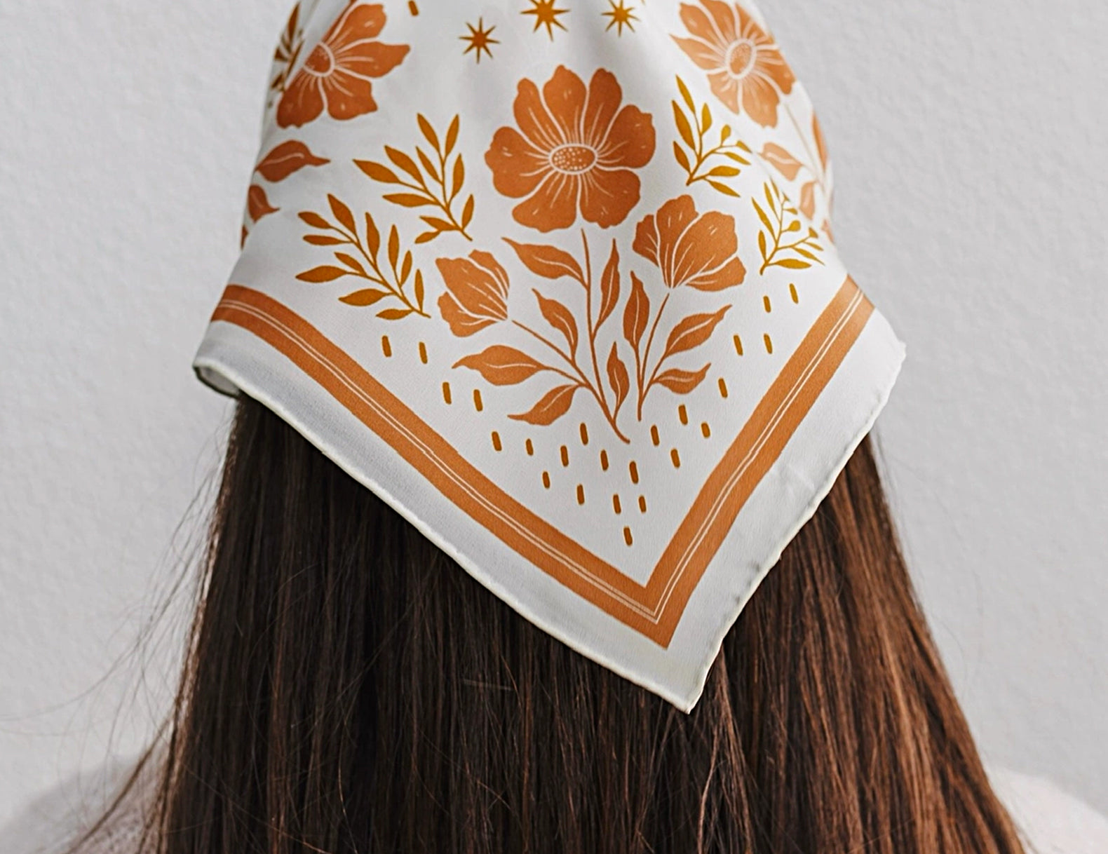 A woman viewed from behind wearing a square white scarf with an orange floral and botanical print, styled over her dark brown hair against a light gray wall.