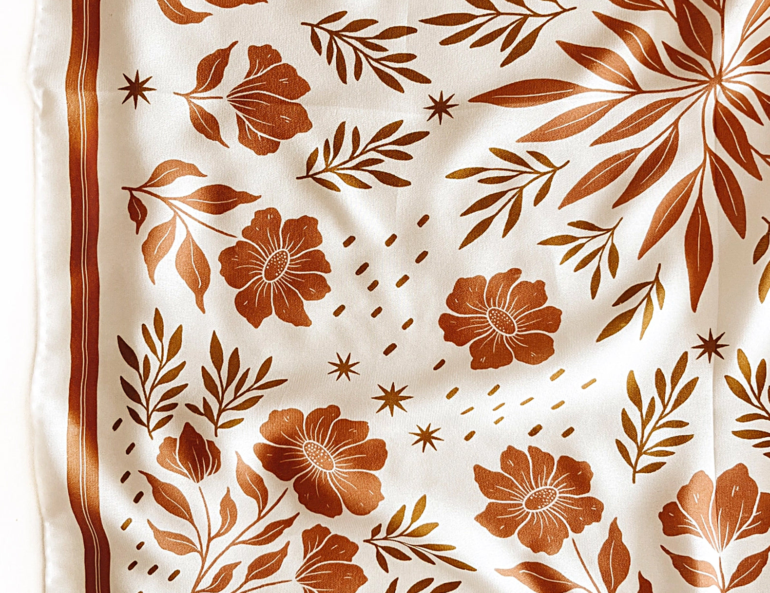 A close-up of a white scarf with a brown floral and botanical pattern, featuring a decorative border and the partial text 'FLORAL WEST' in the bottom right corner.
