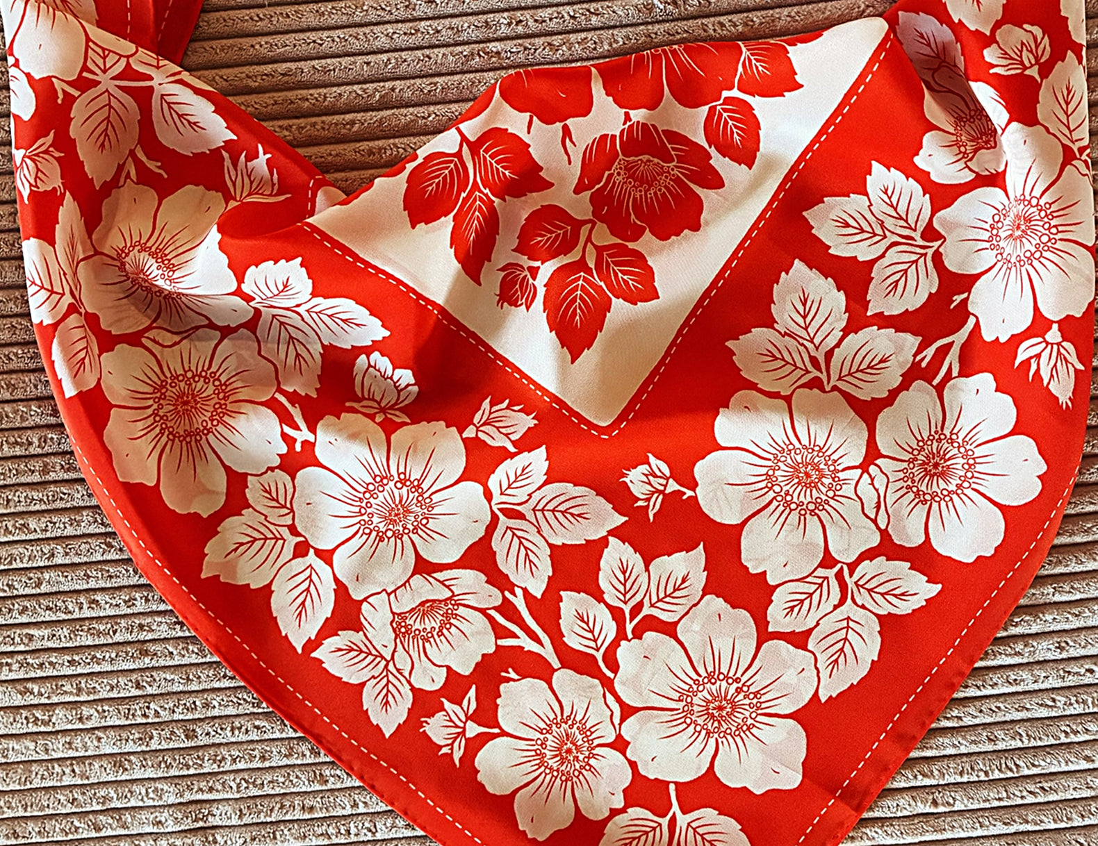 A red and white floral bandana-style pet collar or neck scarf laid flat on a textured beige surface. The fabric features a bold red border and a white background with large red and white cherry blossom-style flowers and leaves.