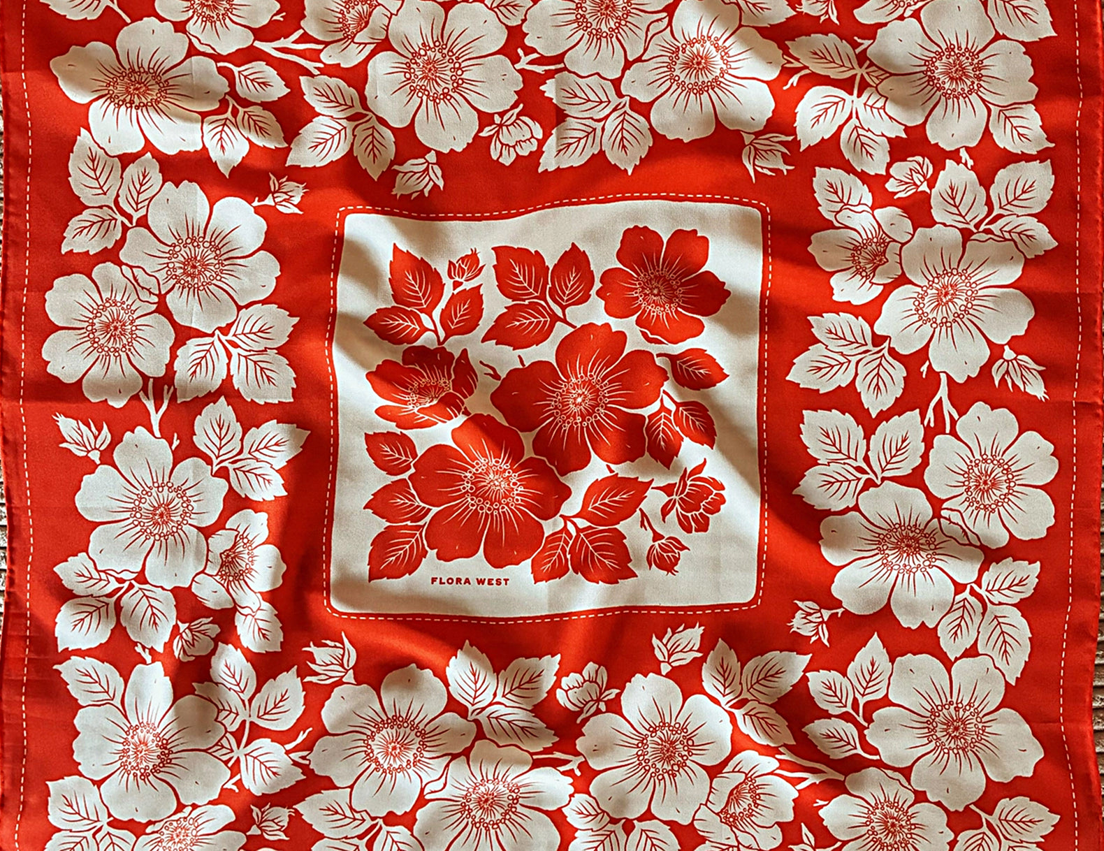 A vintage square silk scarf with a red and white floral pattern, laid flat on a textured beige fabric background. The scarf features a central white square with red flowers and a border of white flowers around the edges.