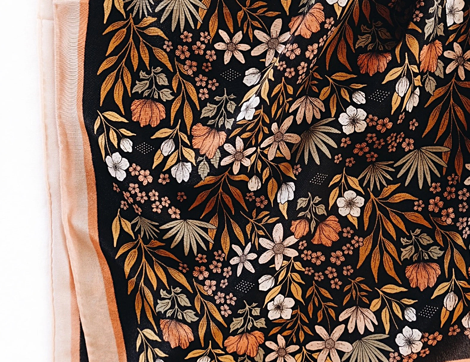 A square scarf with a black background featuring an intricate botanical pattern of orange, brown, and white leaves and flowers, finished with a peach border.