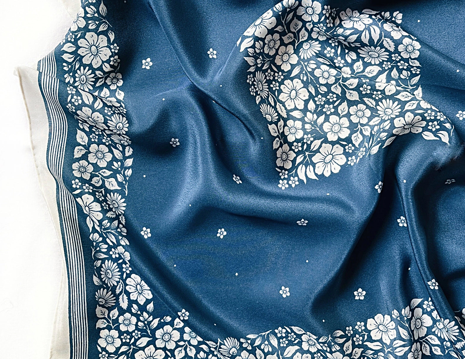 A blue silk scarf with a white floral pattern and striped border, draped elegantly on a white background.
