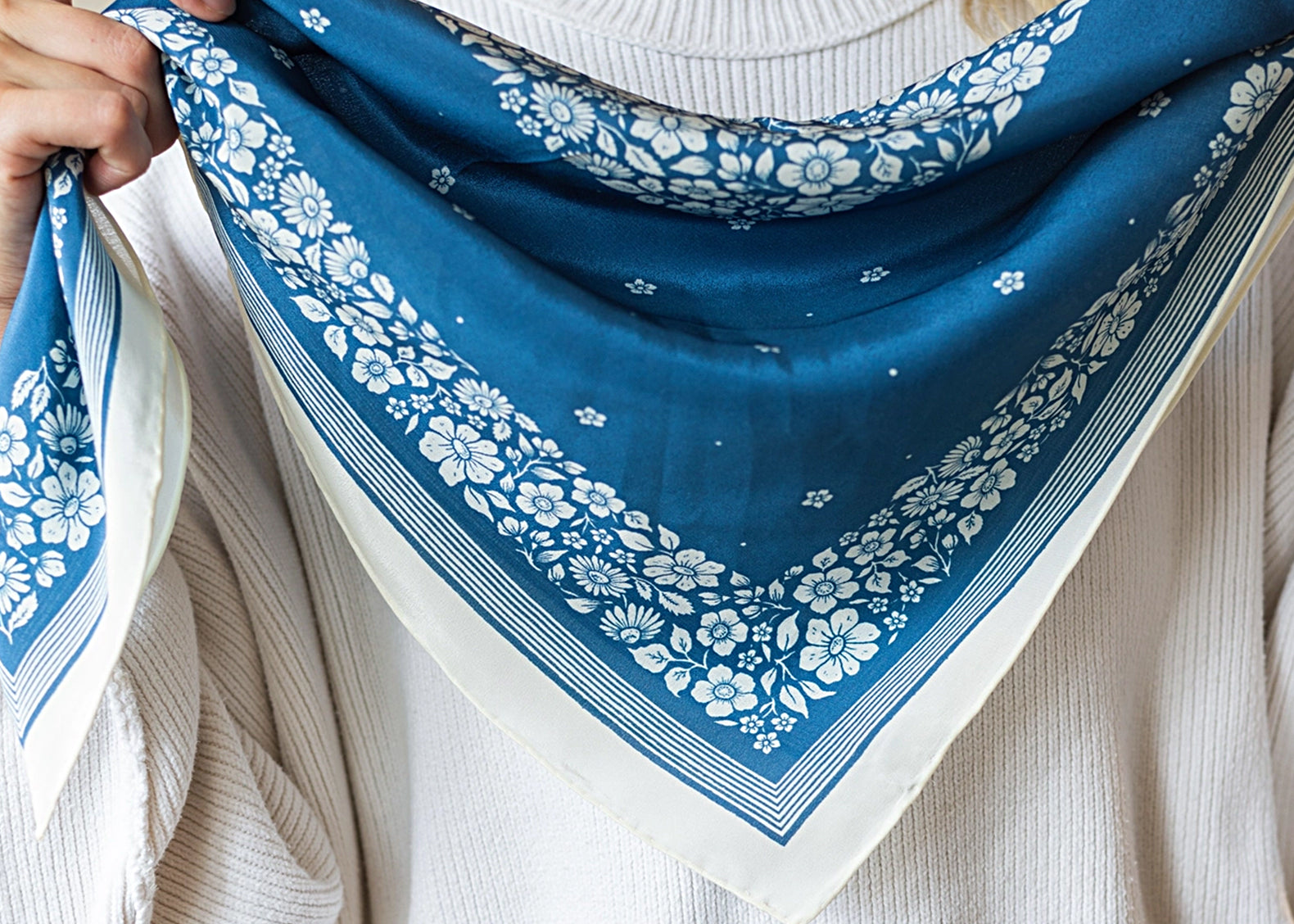 A person wearing a white ribbed sweater holds up a blue bandana-style scarf with a white floral print and border.