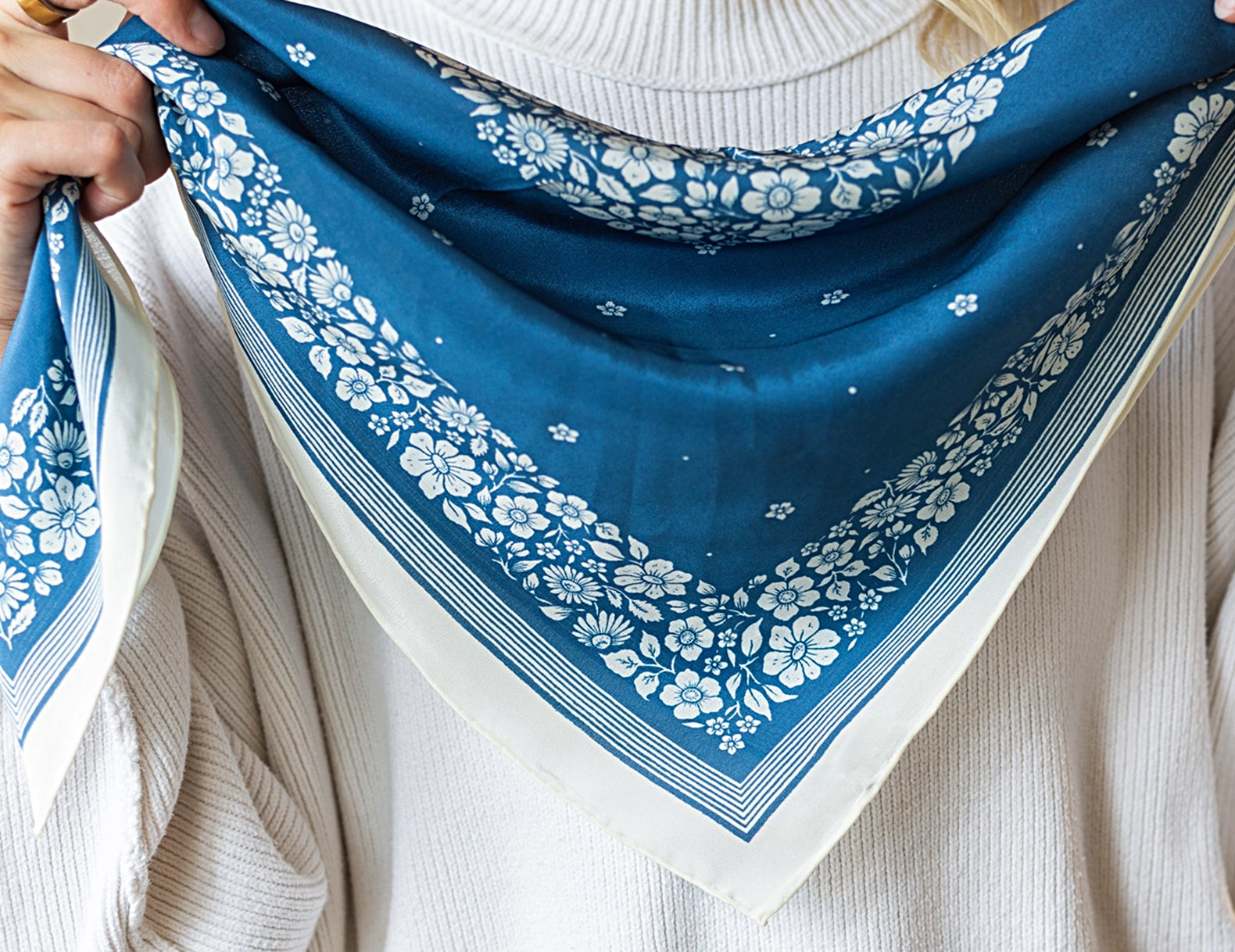 A person wearing a white ribbed sweater holds up a blue bandana-style scarf with a white floral print and border.