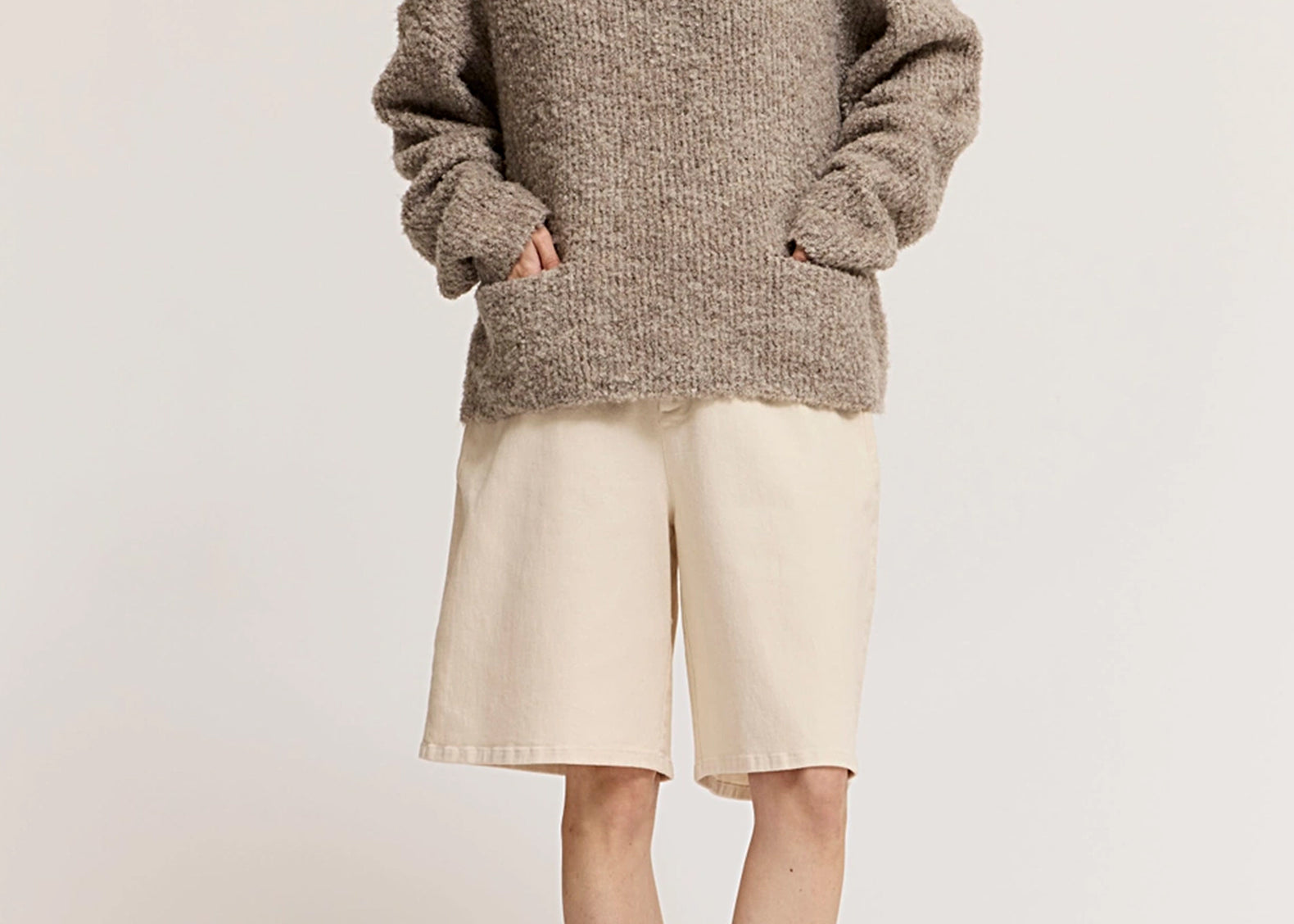 A model wearing a chunky, oversized heathered taupe-brown knit sweater, cream-colored shorts, and dark brown flat shoes, standing against a plain off-white studio background.