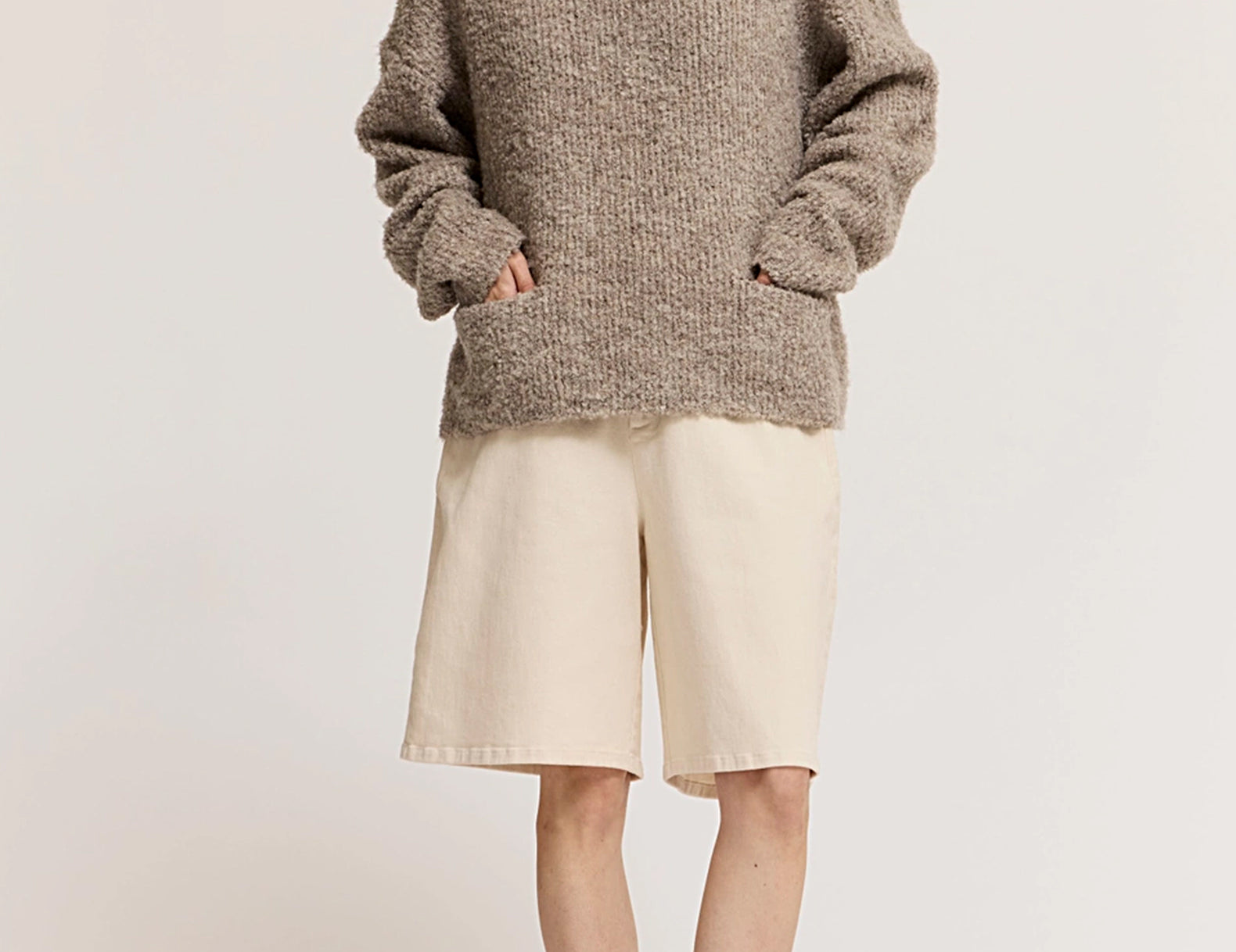 A model wearing a chunky, oversized heathered taupe-brown knit sweater, cream-colored shorts, and dark brown flat shoes, standing against a plain off-white studio background.