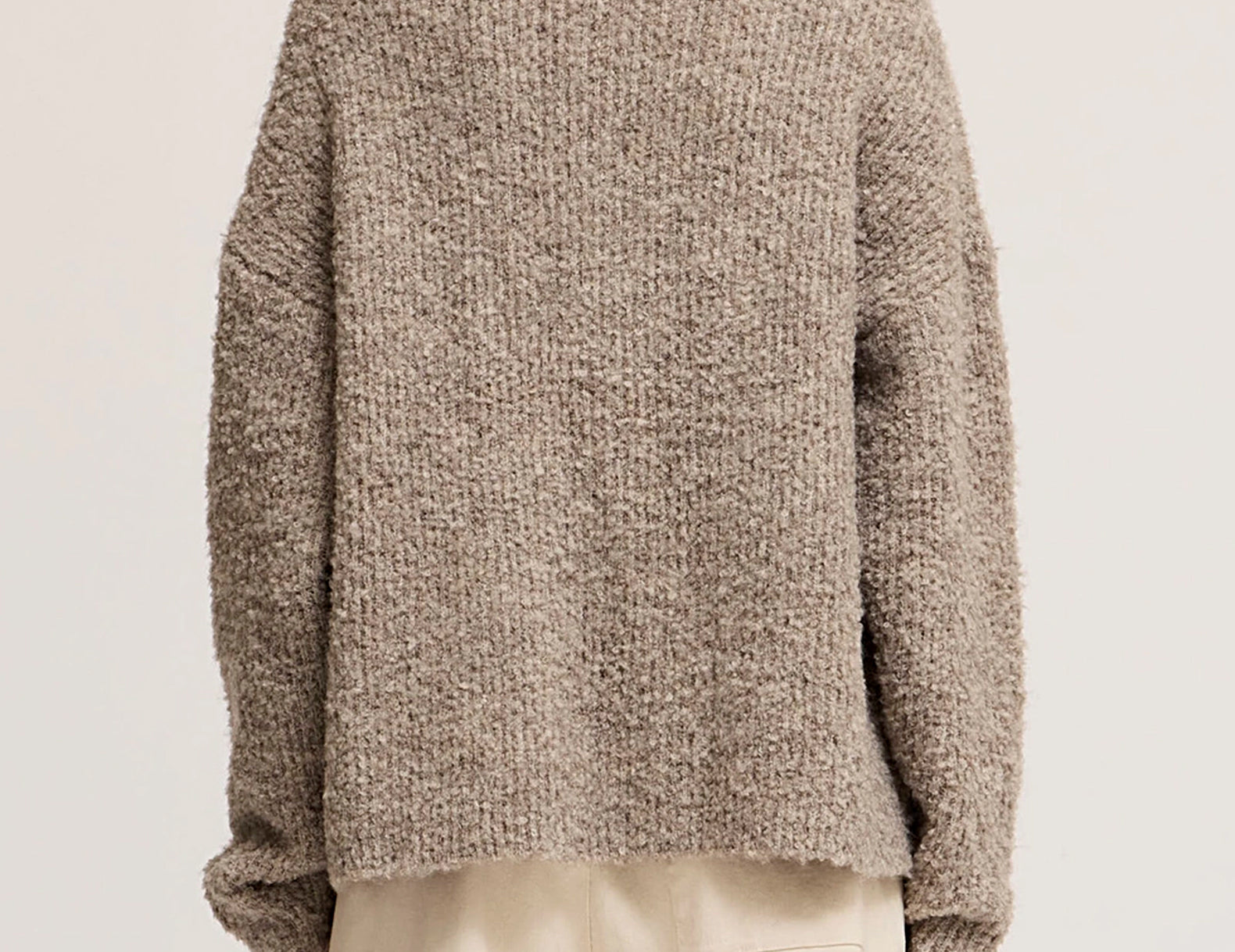 A person shown from the back wearing a chunky knit sweater in a heathered taupe-brown color, paired with light beige pants, against a plain light beige studio background.