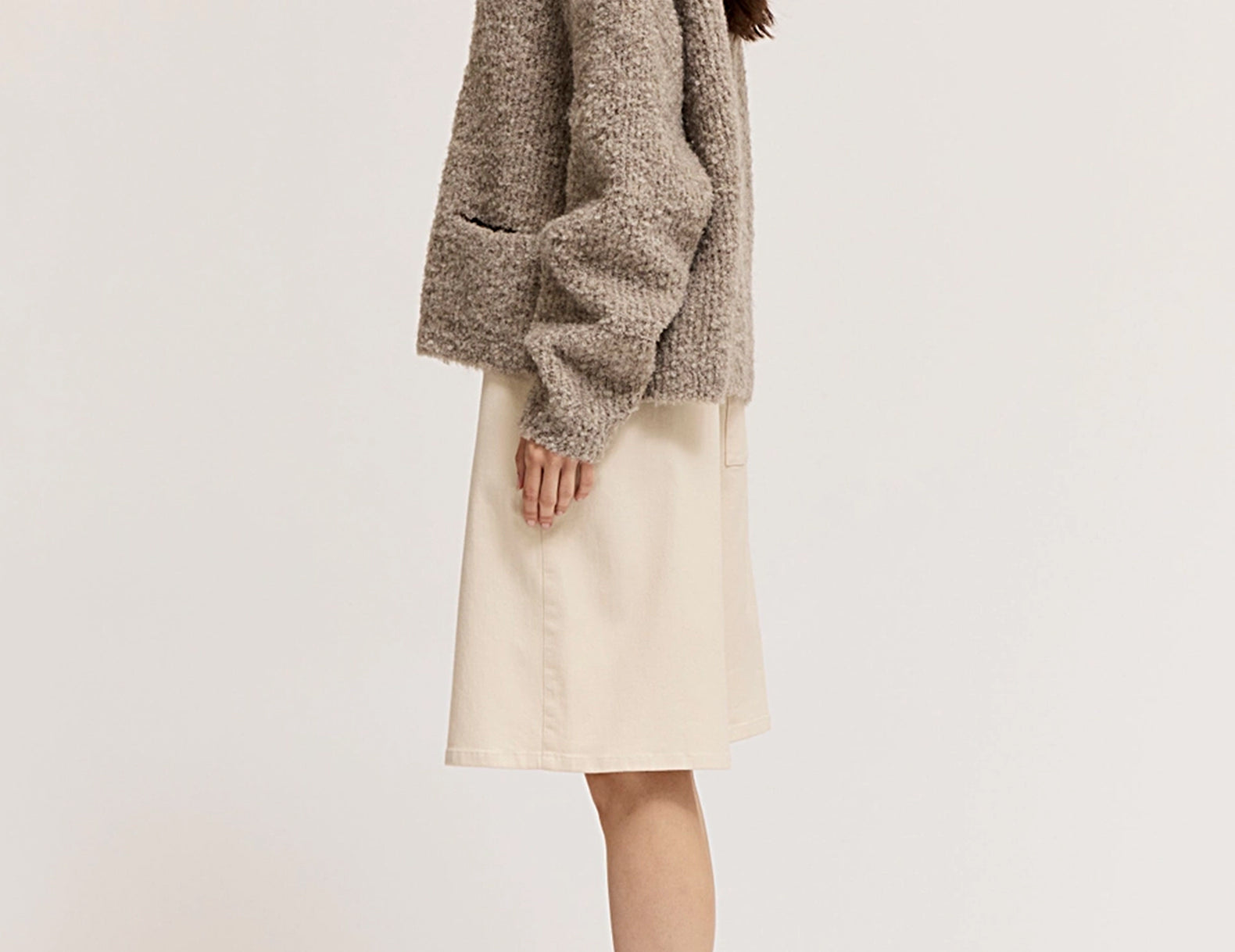 A woman shown in side profile wearing a textured taupe-brown fuzzy cardigan, a light beige knee-length skirt, and dark brown flat shoes against a plain off-white studio background.