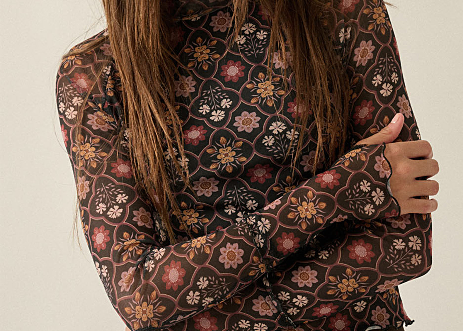 A woman wearing a sheer long-sleeve blouse with an intricate floral and geometric pattern in brown, pink, and beige tones, paired with dark brown trousers, set against a plain light beige background.