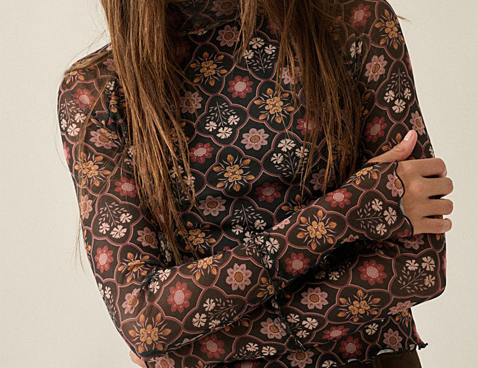 A woman wearing a sheer long-sleeve blouse with an intricate floral and geometric pattern in brown, pink, and beige tones, paired with dark brown trousers, set against a plain light beige background.
