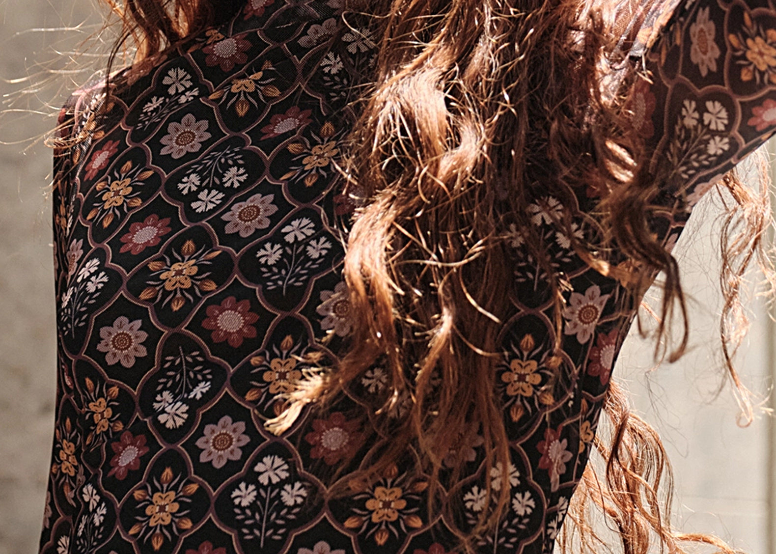 A close-up of a woman wearing a sheer long-sleeve crop top with a bohemian floral pattern in black, rust, pink, and beige. She has long, wavy light brown hair and is wearing gold bracelets.