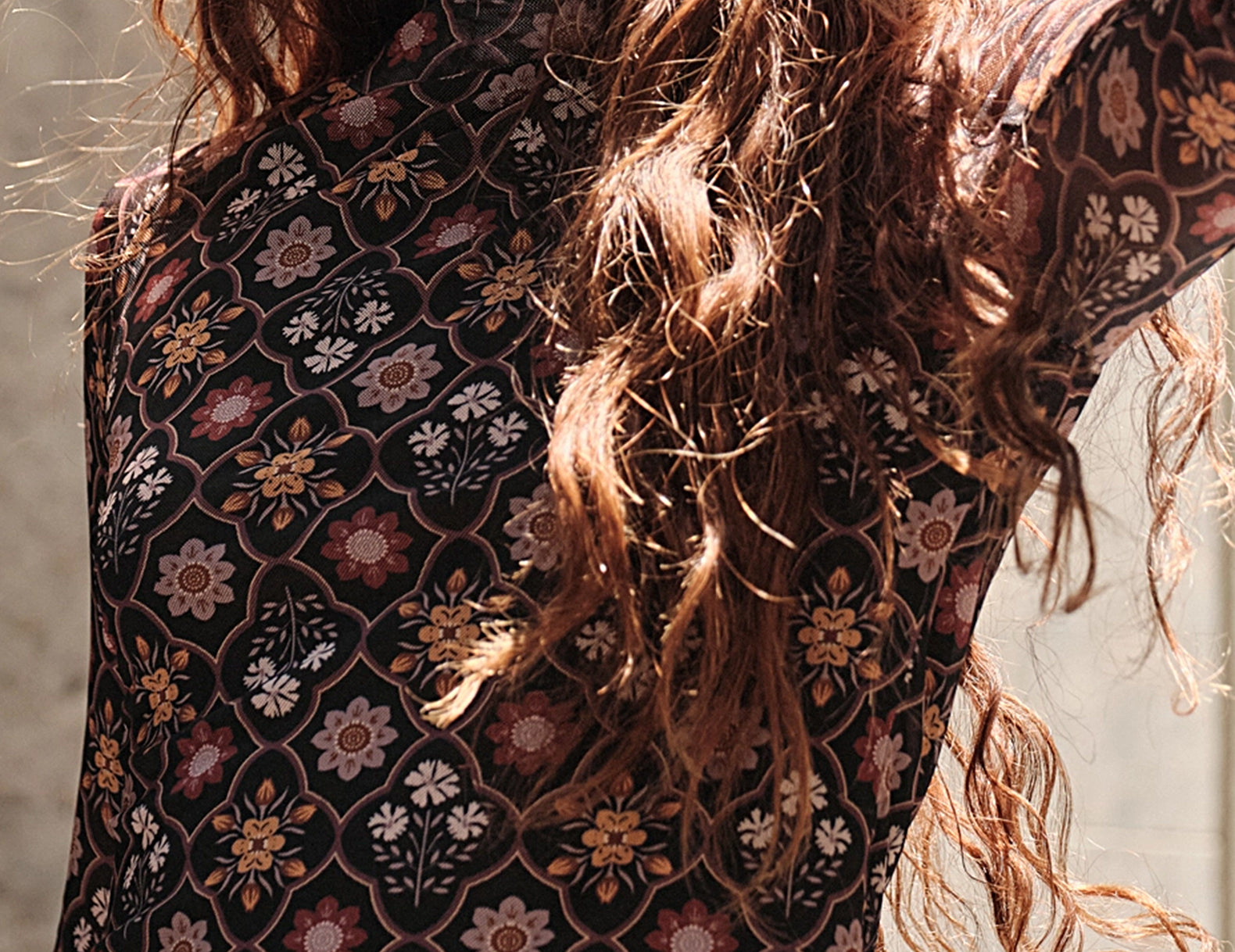 A close-up of a woman wearing a sheer long-sleeve crop top with a bohemian floral pattern in black, rust, pink, and beige. She has long, wavy light brown hair and is wearing gold bracelets.