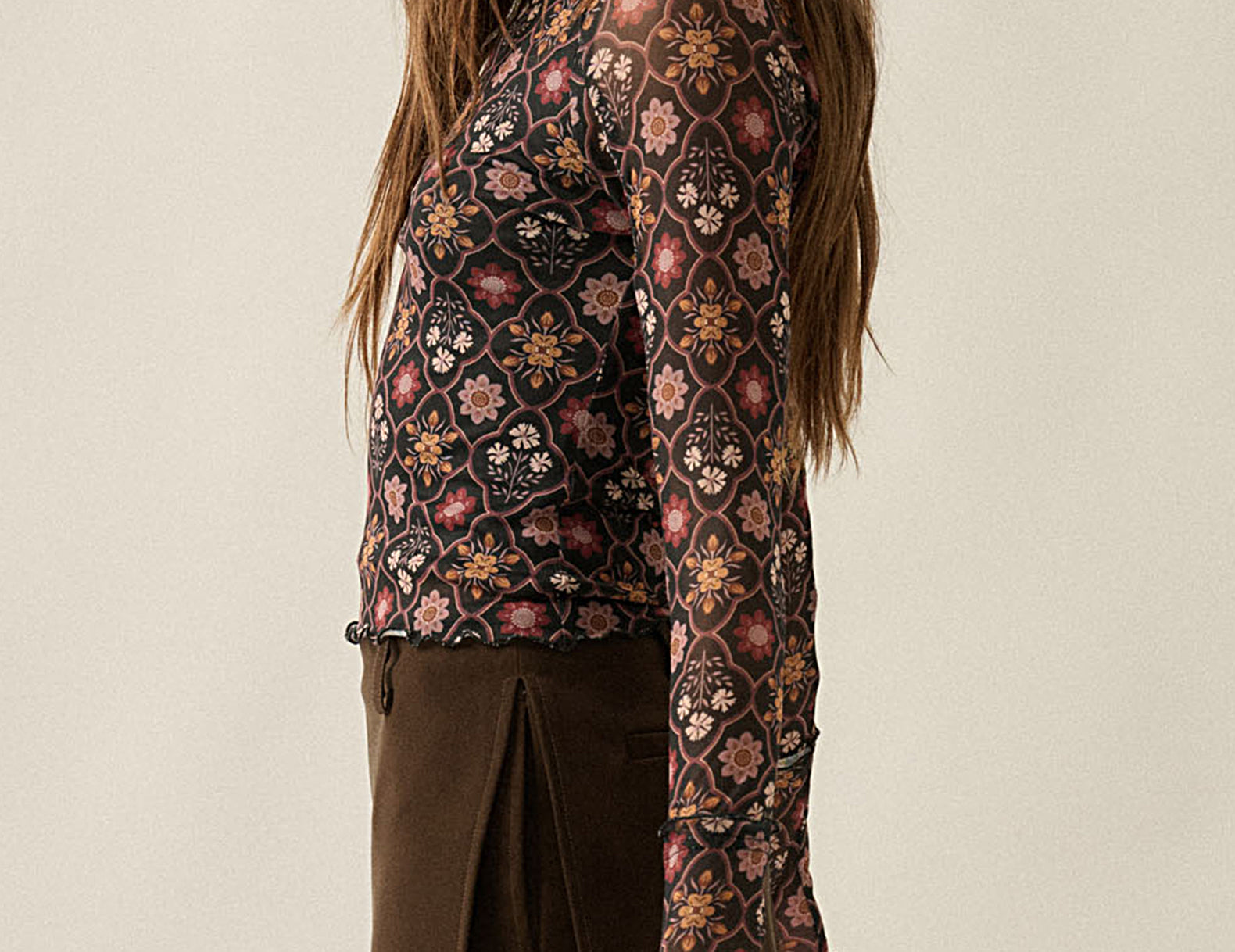 A woman shown from the side wearing a long-sleeve collared blouse with an intricate multicolor floral and geometric pattern in brown, pink, and beige, paired with a brown pleated skirt, against a plain beige background.