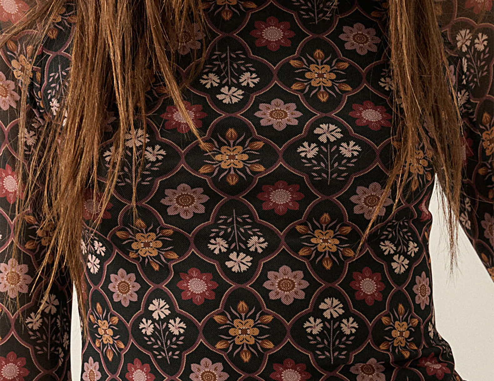 A close-up of a woman wearing a sheer long-sleeve mock-neck top with a dark brown and black background and a detailed floral pattern in muted pink, beige, and gold.