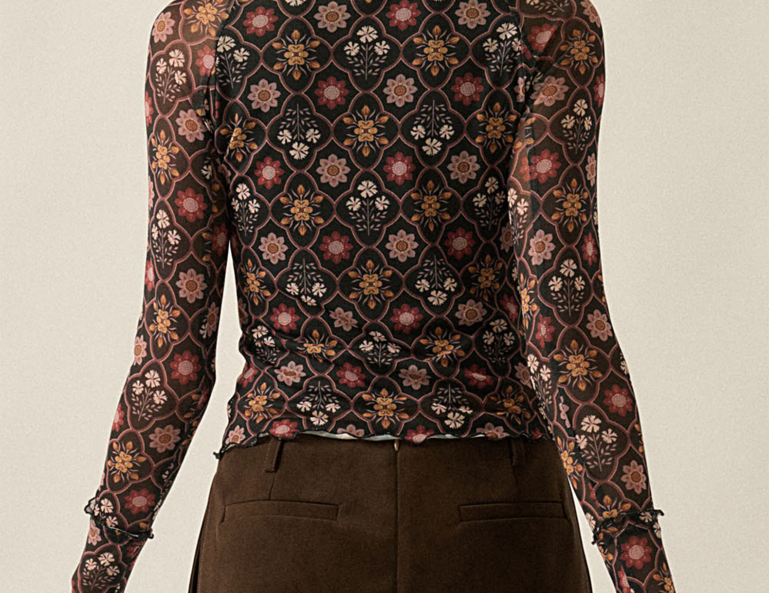 A woman shown from the back wearing a fitted long-sleeve top with an intricate multicolor floral and geometric pattern on a dark background, paired with a brown mini skirt, against a plain light beige studio backdrop.