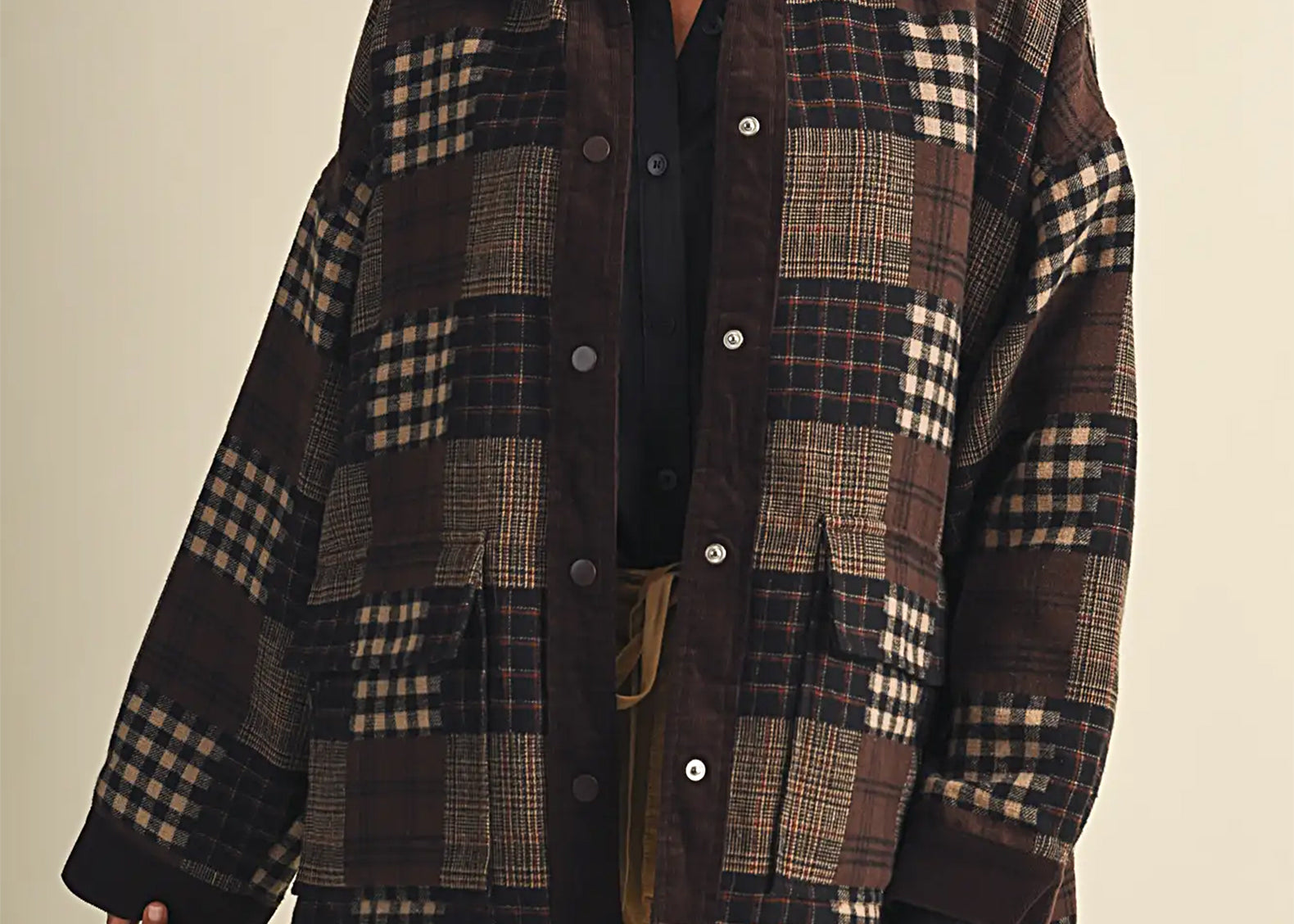 A woman wearing a dark brown and beige patchwork plaid button-up shirt jacket over a black shirt and tan pants, photographed against a solid light beige background.