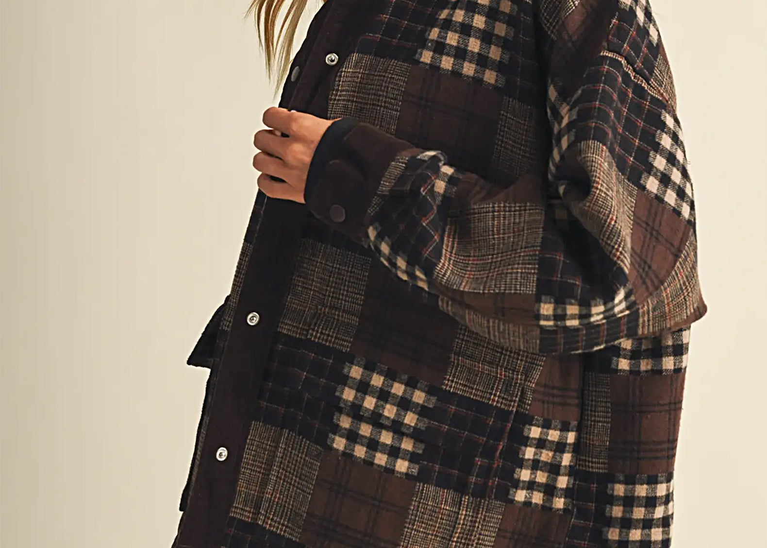A woman wearing a dark brown and beige patchwork plaid button-up shirt jacket, paired with tan cargo pants, photographed against a solid light beige background.