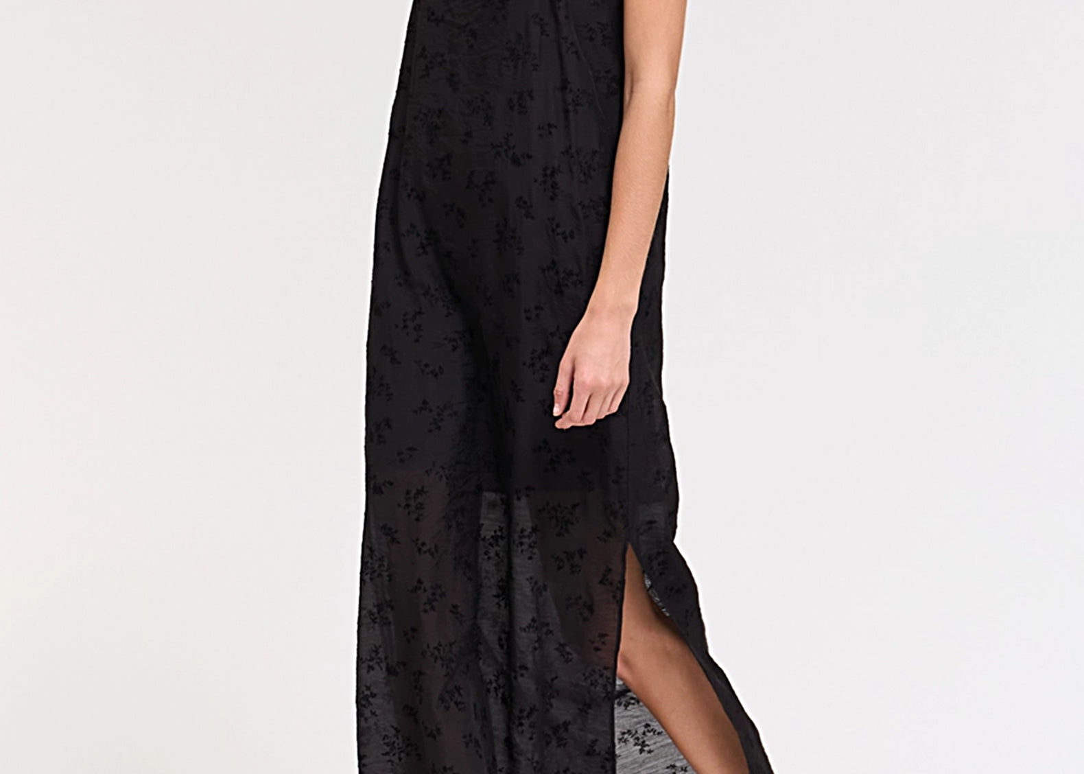 A woman modeling a black sheer floral maxi dress with spaghetti straps and a high side slit, paired with brown pointed-toe heels, against a plain white studio background.