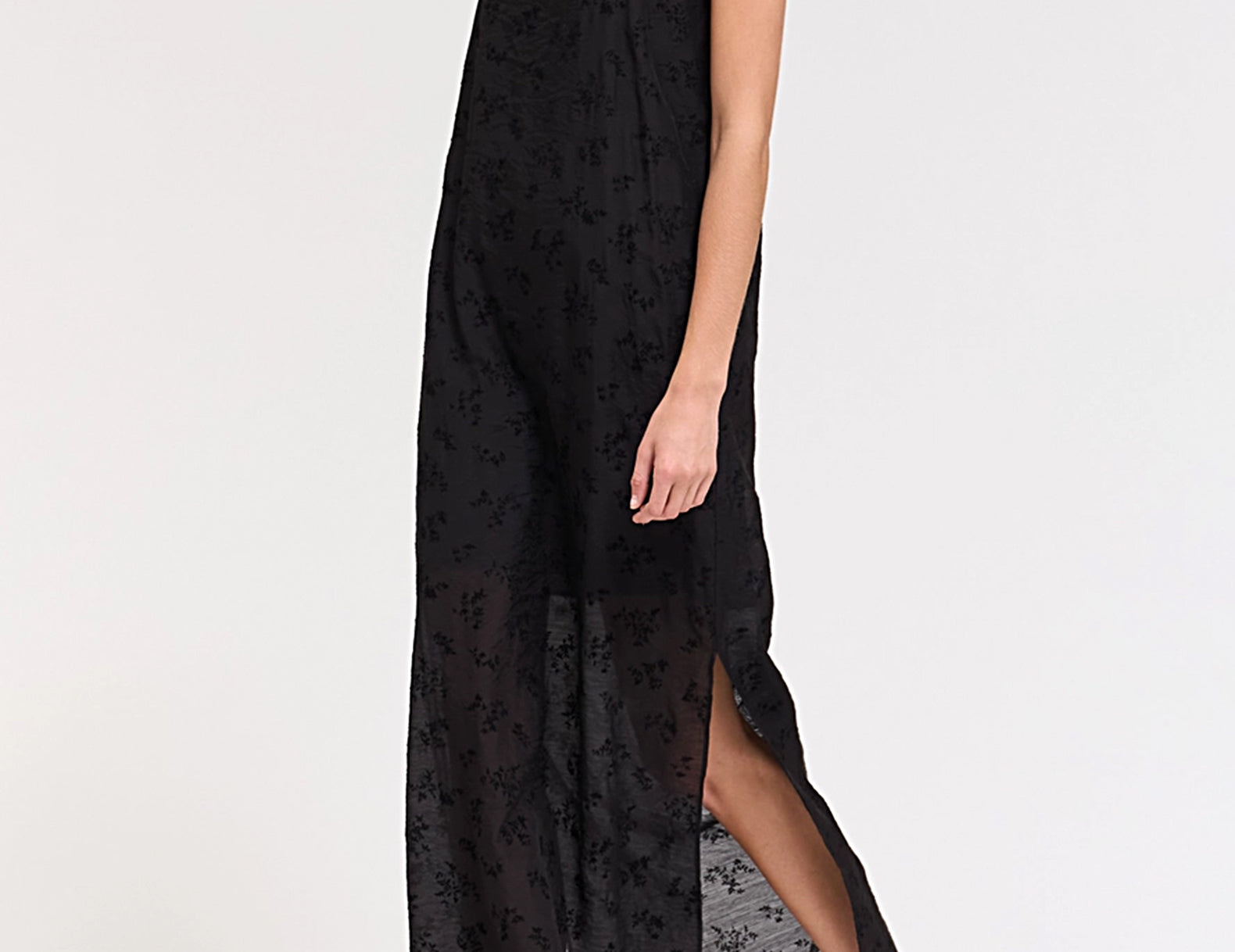 A woman modeling a black sheer floral maxi dress with spaghetti straps and a high side slit, paired with brown pointed-toe heels, against a plain white studio background.