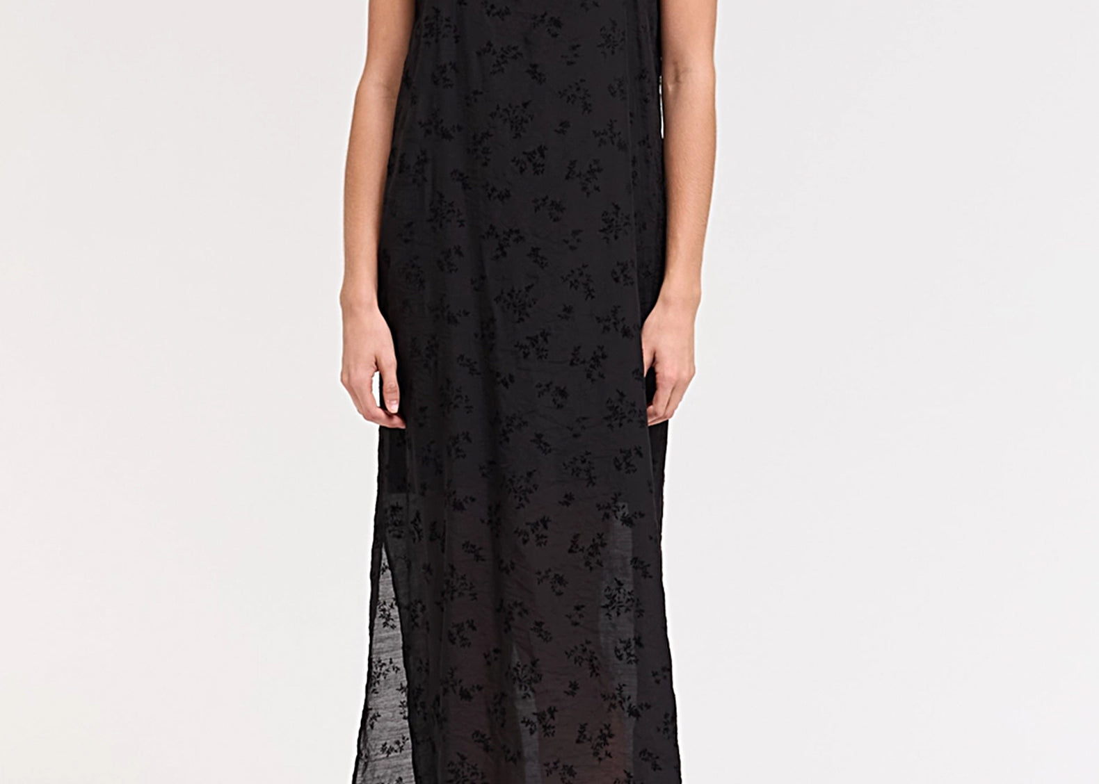 A woman modeling a black spaghetti-strap maxi dress with a sheer floral lace overlay, photographed against a plain white studio background.