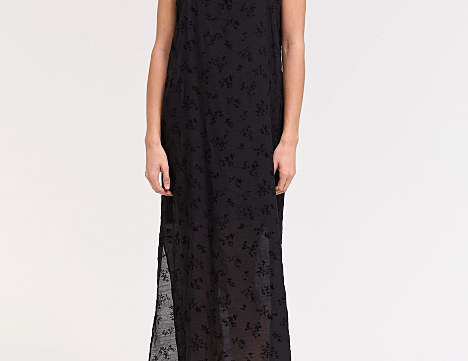 A woman modeling a black spaghetti-strap maxi dress with a sheer floral lace overlay, photographed against a plain white studio background.