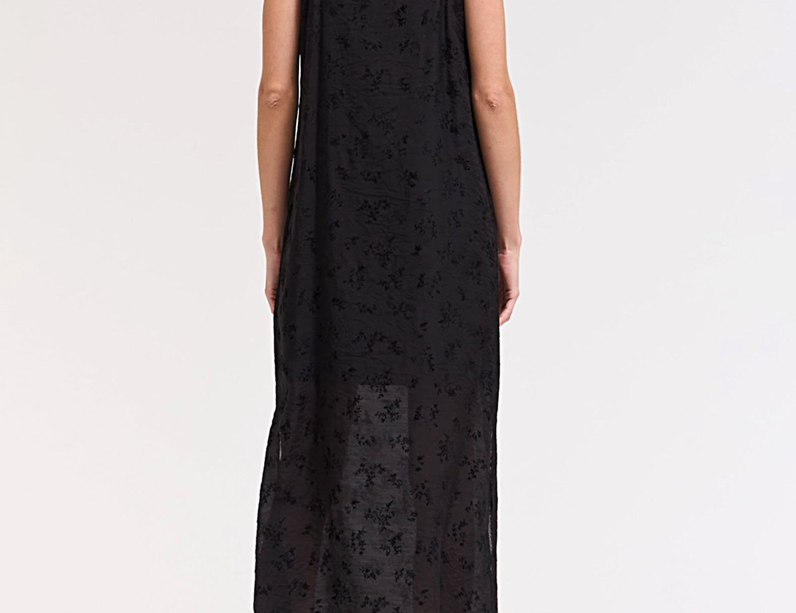 A woman viewed from the back wearing a black spaghetti-strap maxi dress with a sheer floral lace overlay, paired with black heeled sandals, against a plain white studio background.