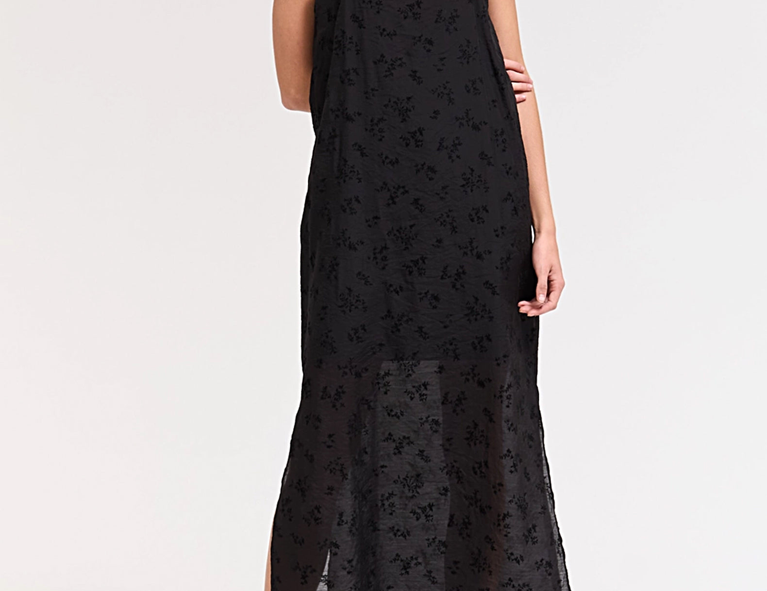 A woman modeling a black spaghetti-strap maxi dress with a subtle tonal floral pattern and a sheer overlay, paired with brown pointed-toe heels against a plain white studio background.