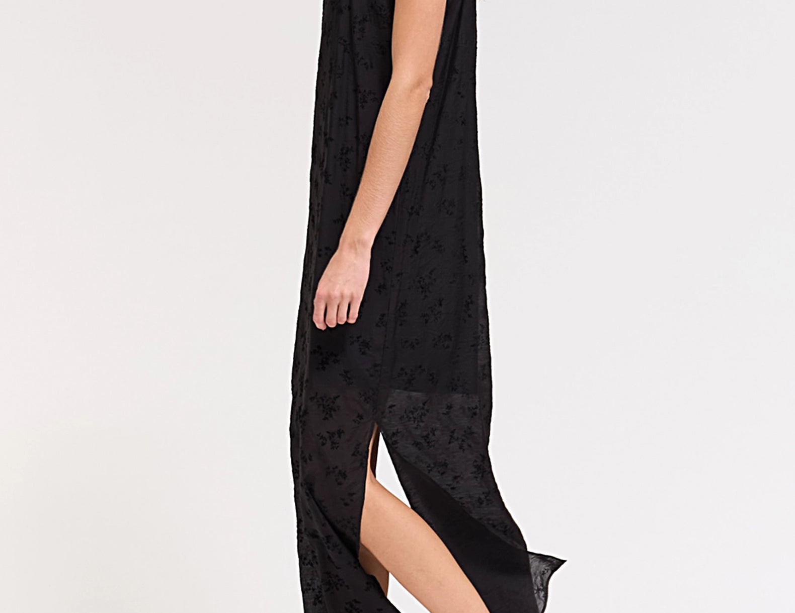A woman modeling a black sleeveless maxi dress with a subtle tonal floral pattern, shown from a side profile against a clean white studio background.