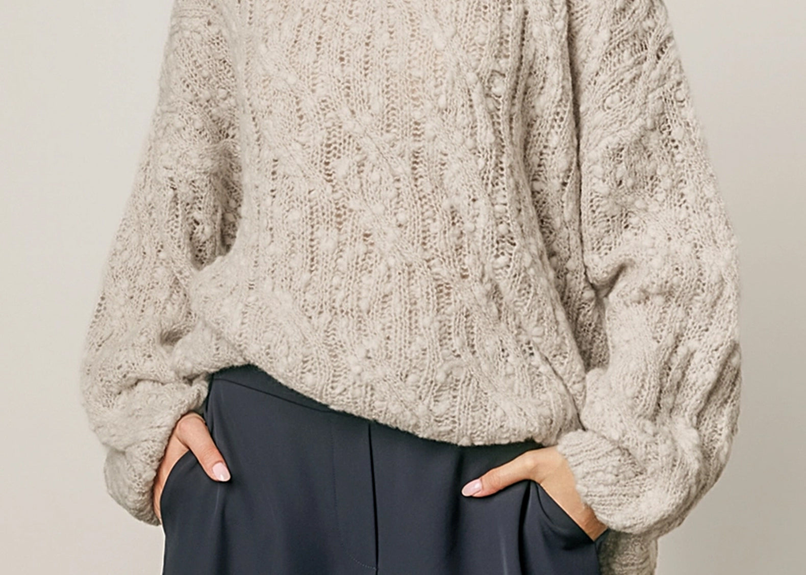 A woman wearing a chunky, textured knit sweater in a light taupe-beige color, paired with dark navy wide-leg trousers, against a plain light gray background.