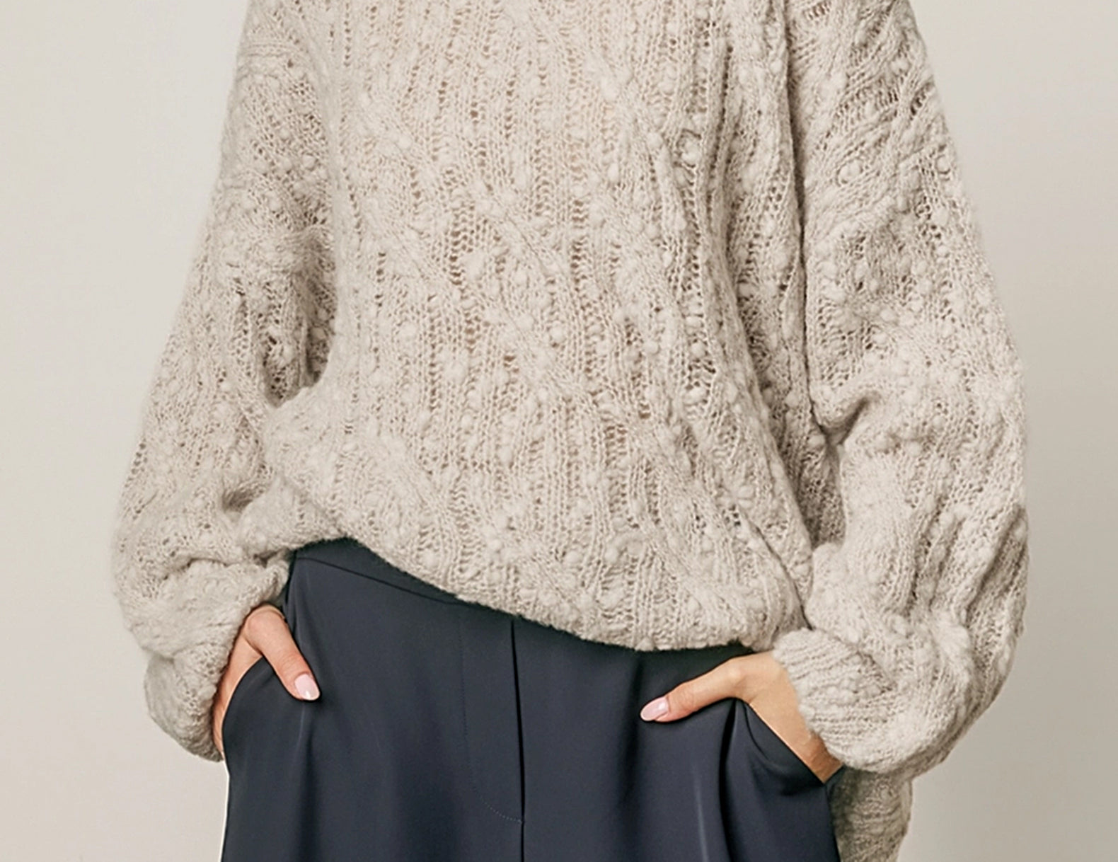 A woman wearing a chunky, textured knit sweater in a light taupe-beige color, paired with dark navy wide-leg trousers, against a plain light gray background.