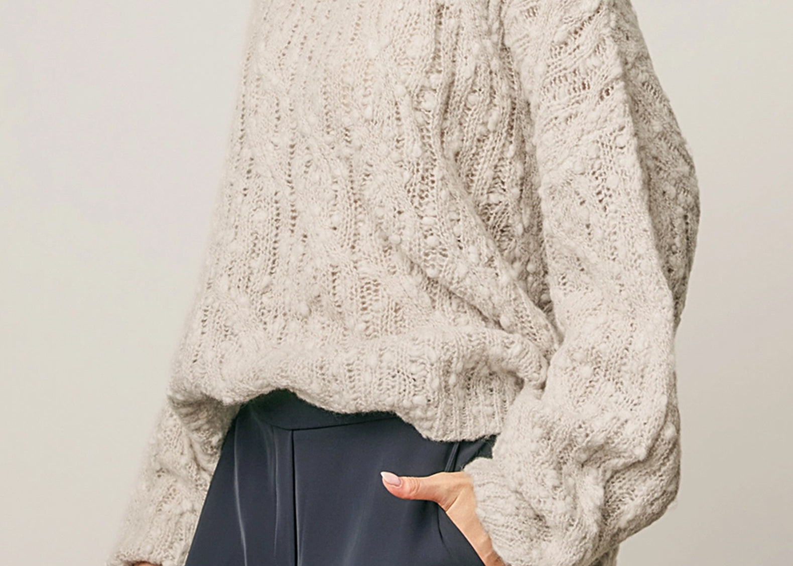A woman wearing a chunky, textured cream-colored knit sweater with balloon sleeves, paired with dark slate wide-leg trousers, against a neutral light gray background.