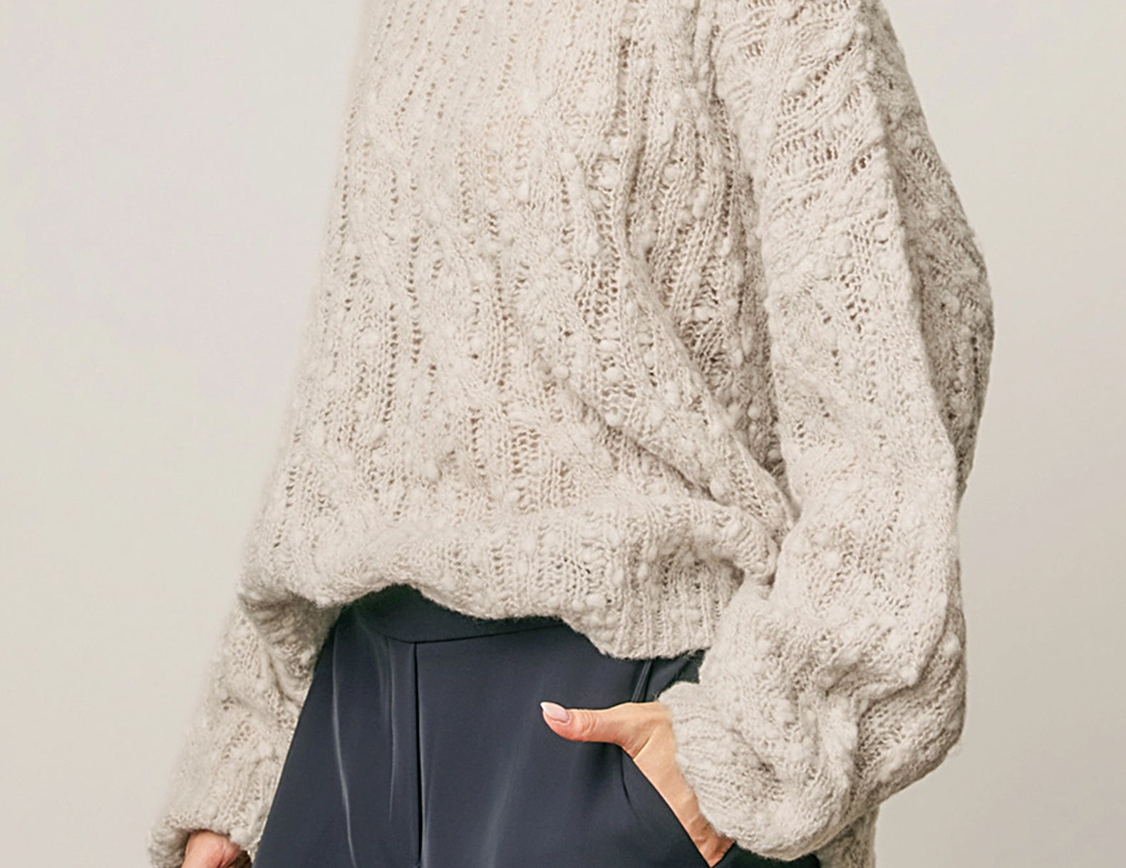 A woman wearing a chunky, textured cream-colored knit sweater with balloon sleeves, paired with dark slate wide-leg trousers, against a neutral light gray background.