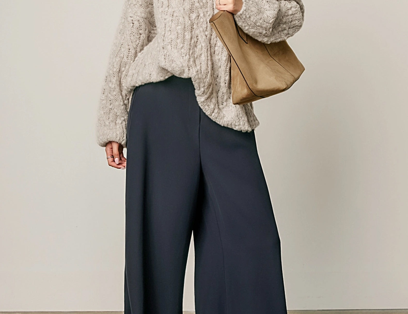 A model wearing a chunky light beige knit sweater, dark navy wide-leg trousers, and carrying a tan leather shoulder bag against a neutral studio background.