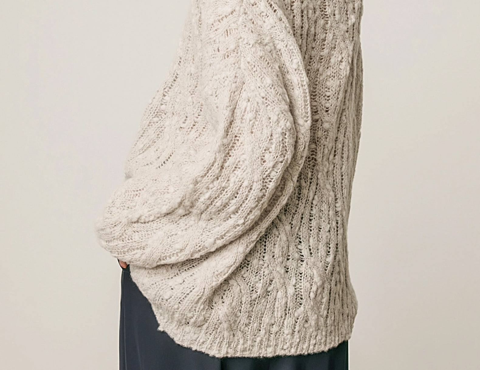 A woman wearing a chunky, textured knit sweater in a light oatmeal beige color, paired with dark navy wide-leg trousers, shown from a side-back angle against a plain light gray studio background.
