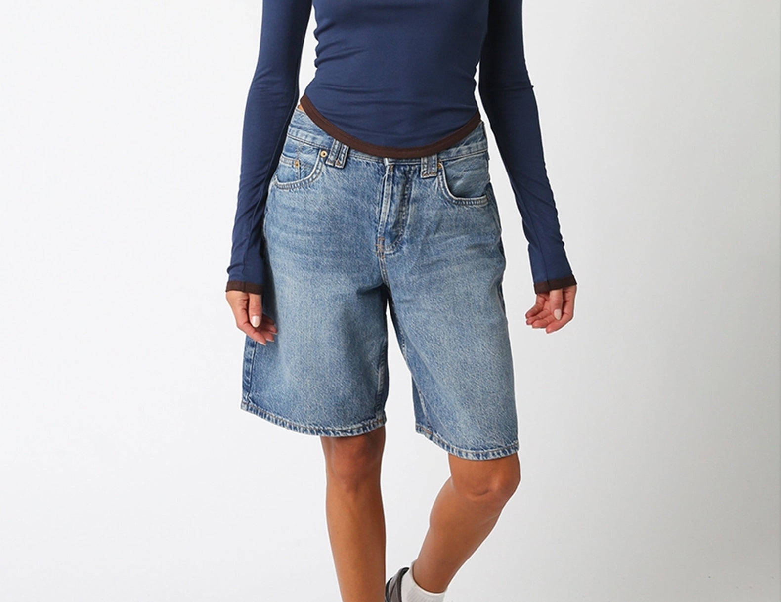A model wearing a navy blue long-sleeve top, light-wash denim shorts, white socks, and brown leather loafers, standing against a plain white studio background.