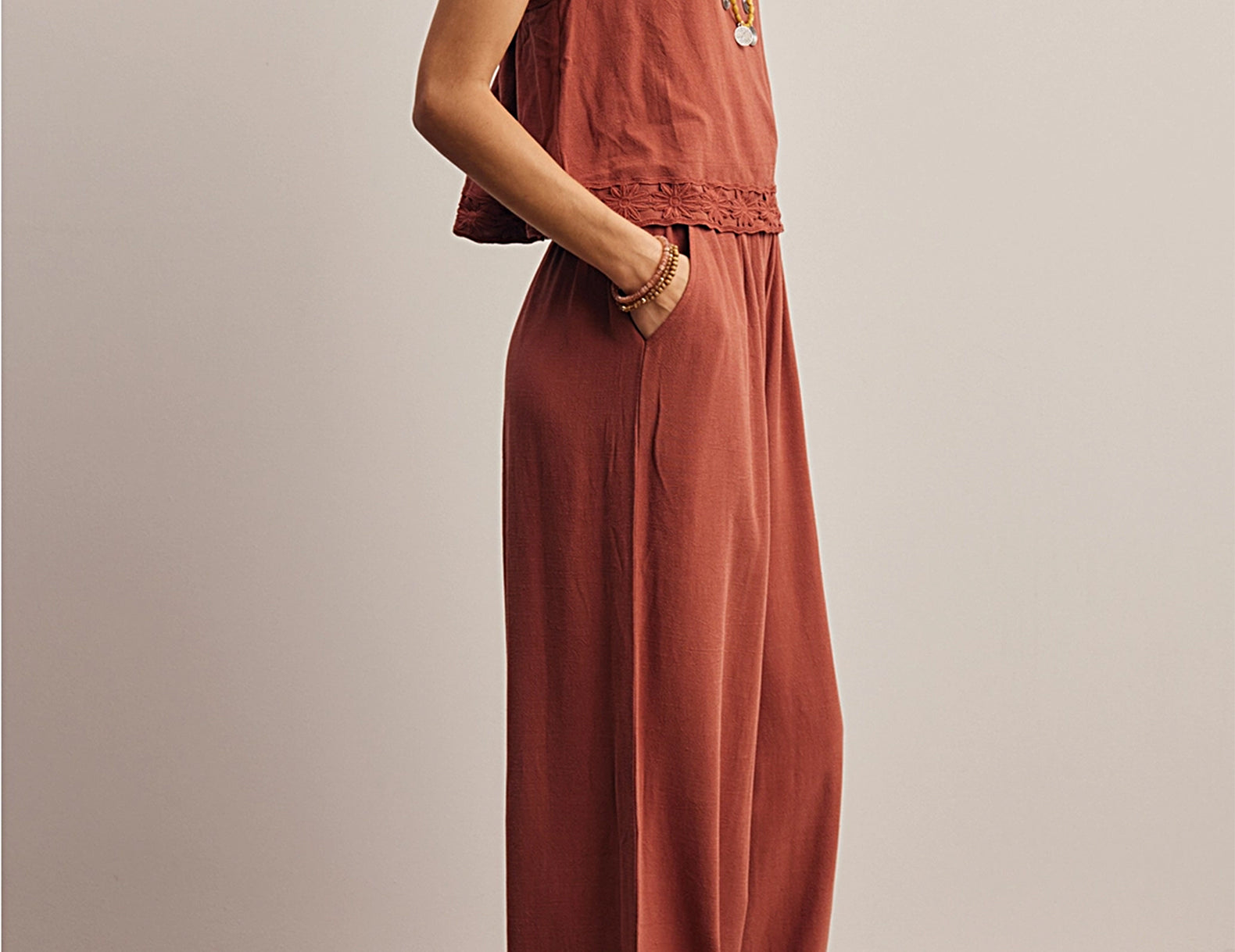 A woman models a rust-colored sleeveless jumpsuit with lace-up shoulder straps, delicate lace trim at the neckline and hem, and wide-leg pants, set against a neutral studio background.