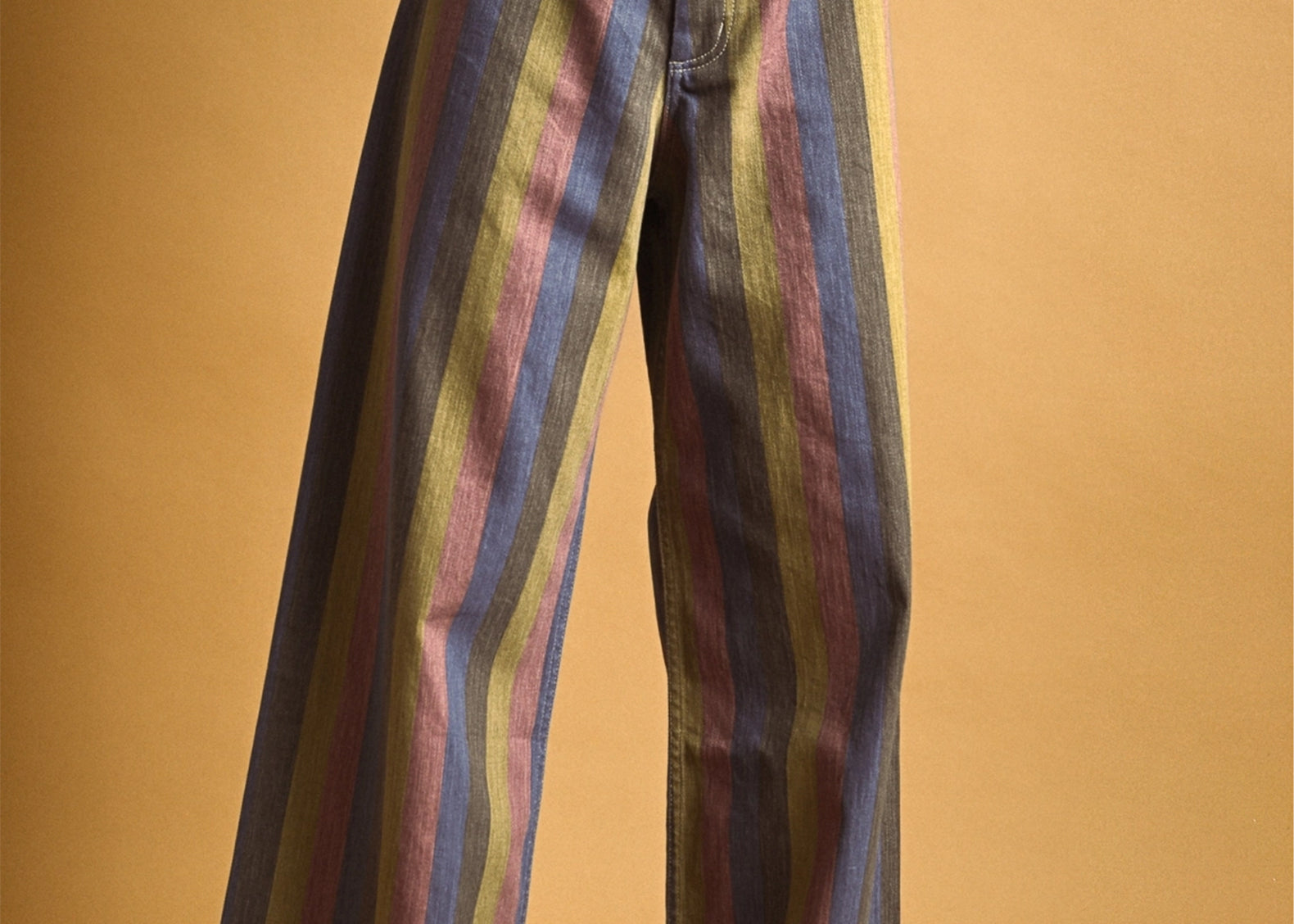 A person wearing wide-leg striped pants with vertical stripes in blue, purple, green, and yellow, paired with a red top and tan shoes, against a solid tan studio background.