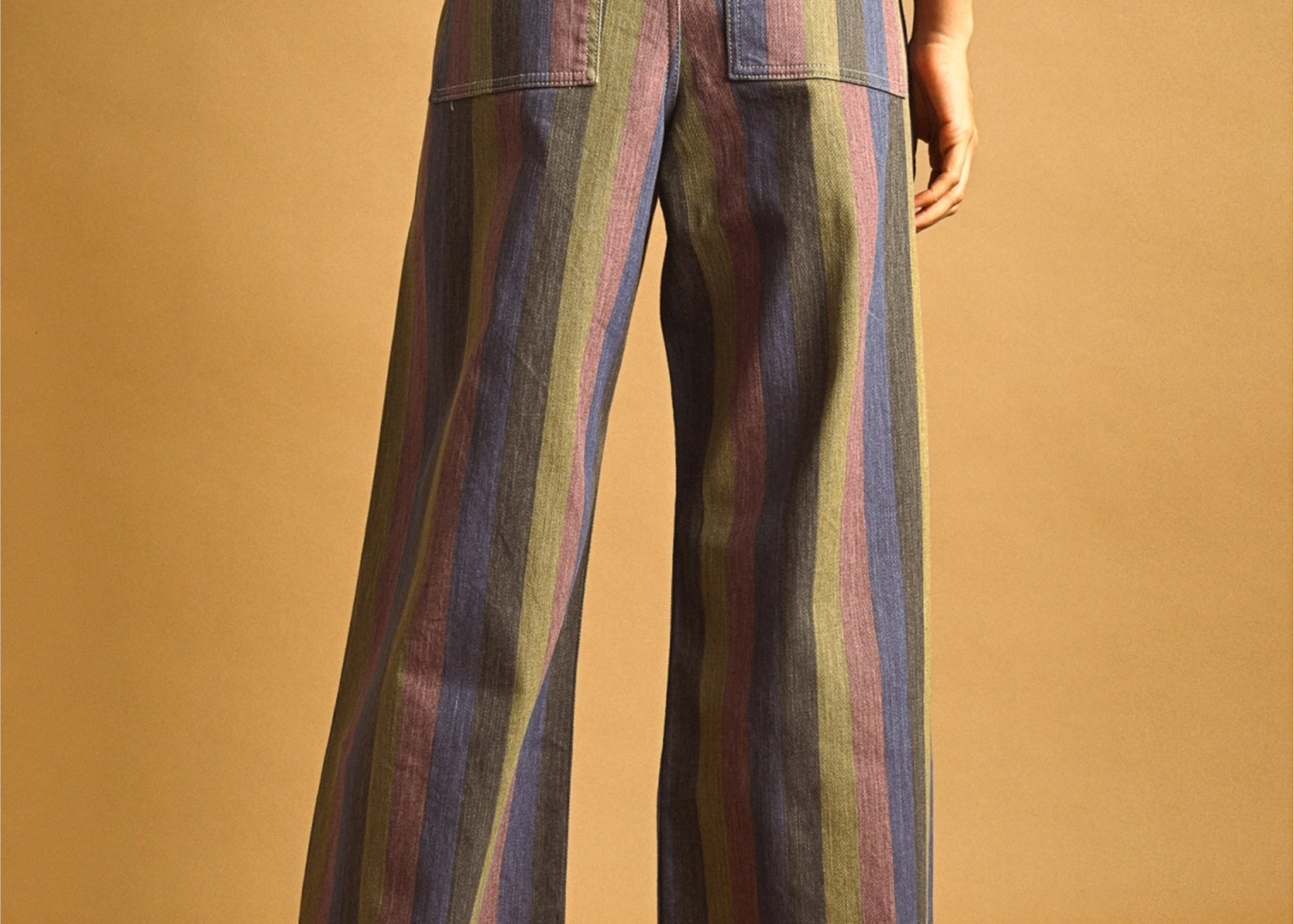 A model shown from the back wearing wide-leg trousers with vertical stripes in muted blue, green, purple, and pink, paired with a red top against a solid tan studio background.