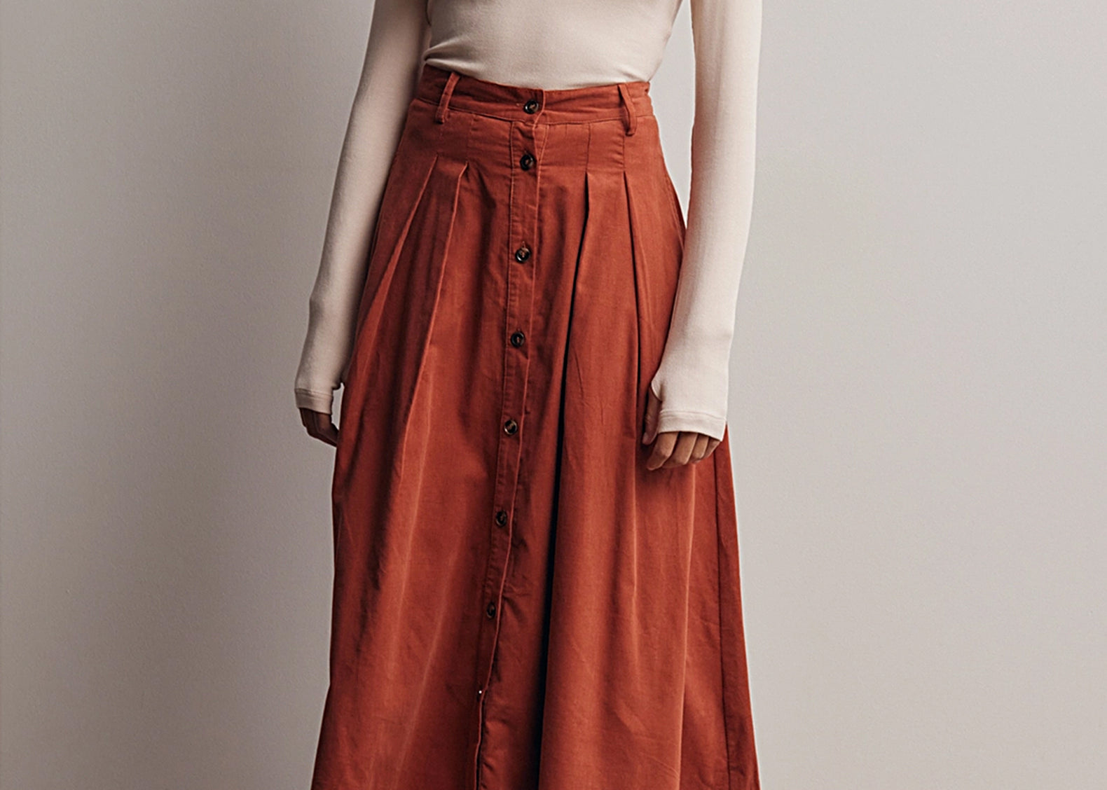 Front view of the Marnie Pleated Button Front Corduroy Skirt, a terracotta A-line midi worn with a cream fitted turtleneck