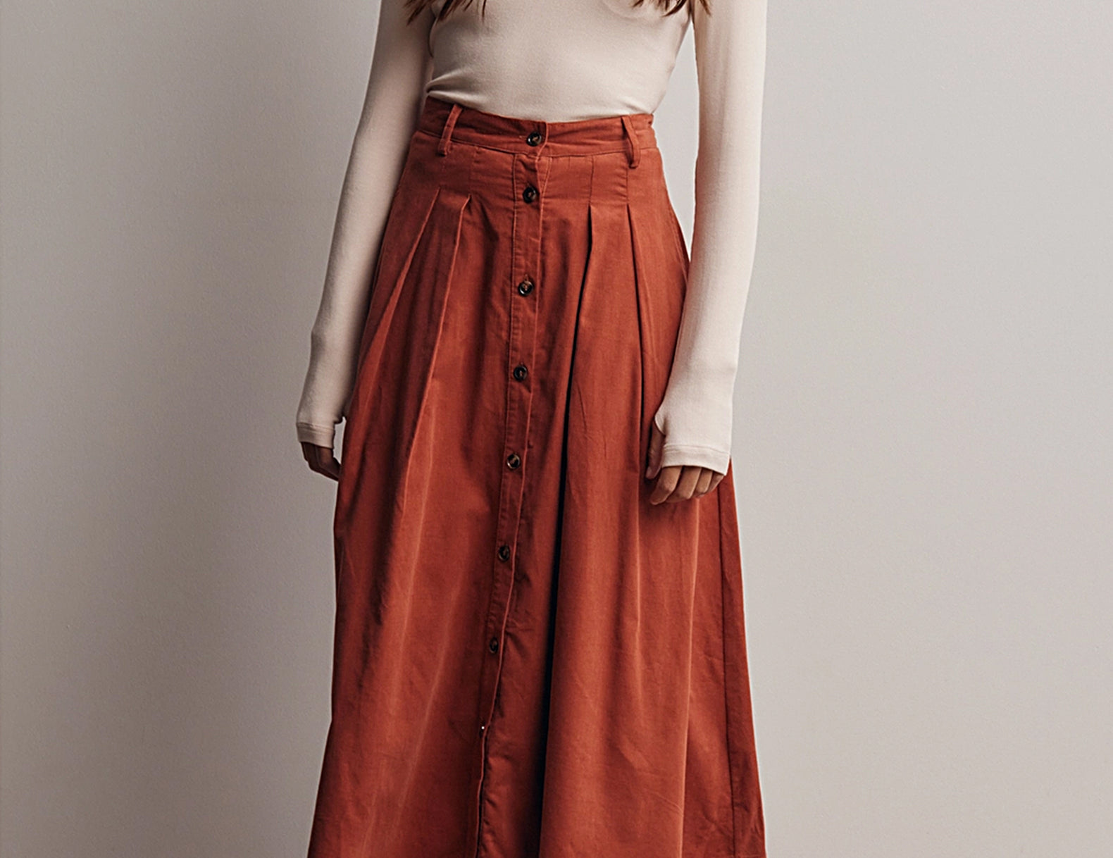 Front view of the Marnie Pleated Button Front Corduroy Skirt, a terracotta A-line midi worn with a cream fitted turtleneck