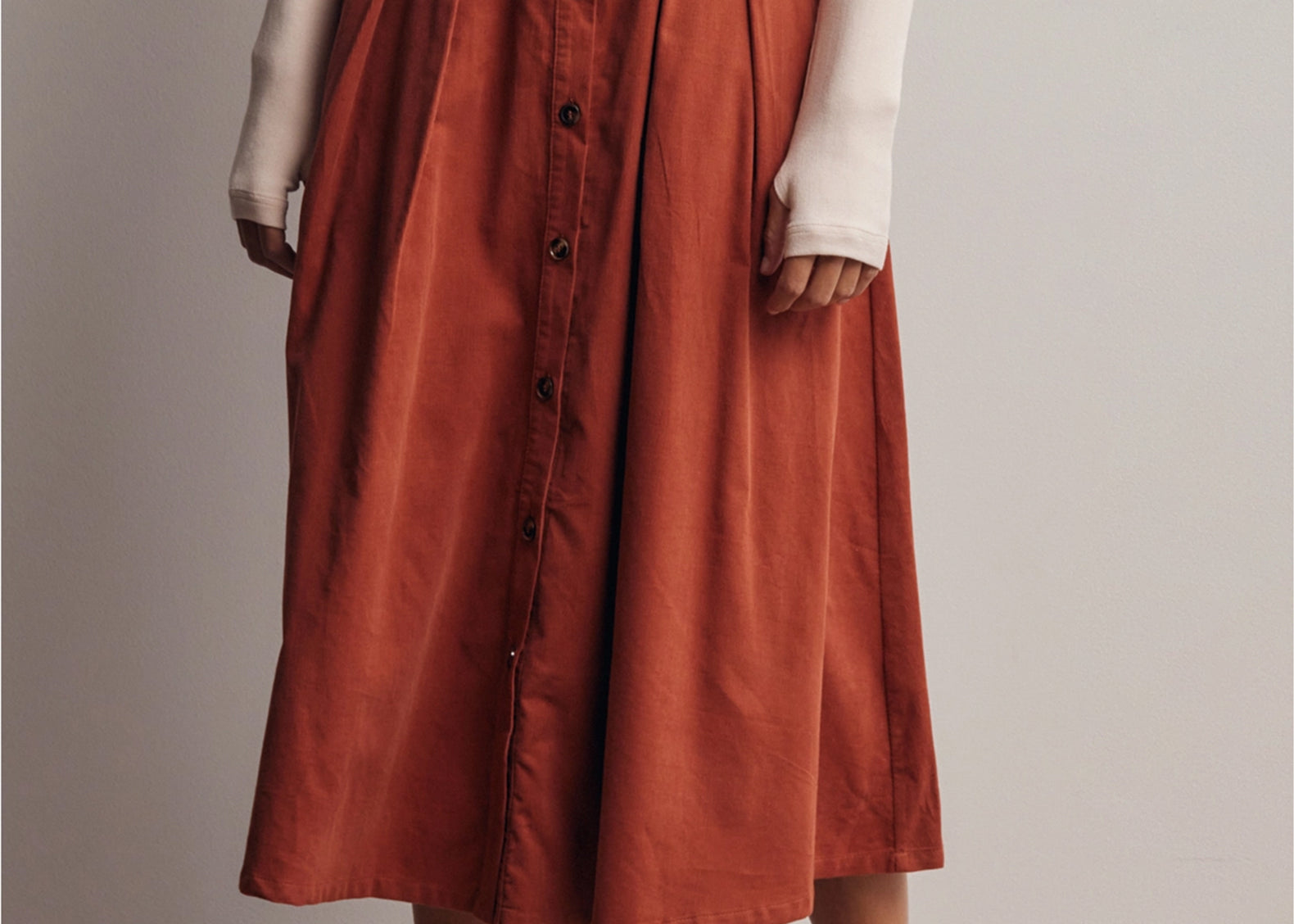 A model wearing a rust-orange high-waisted midi skirt with a button-down front, styled with a cream long-sleeve top and beige loafers against a neutral studio background.