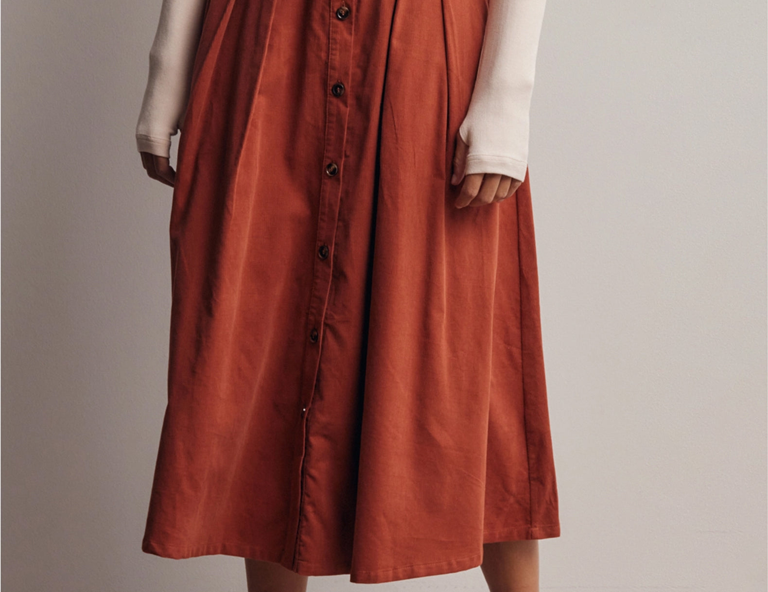 A model wearing a rust-orange high-waisted midi skirt with a button-down front, styled with a cream long-sleeve top and beige loafers against a neutral studio background.