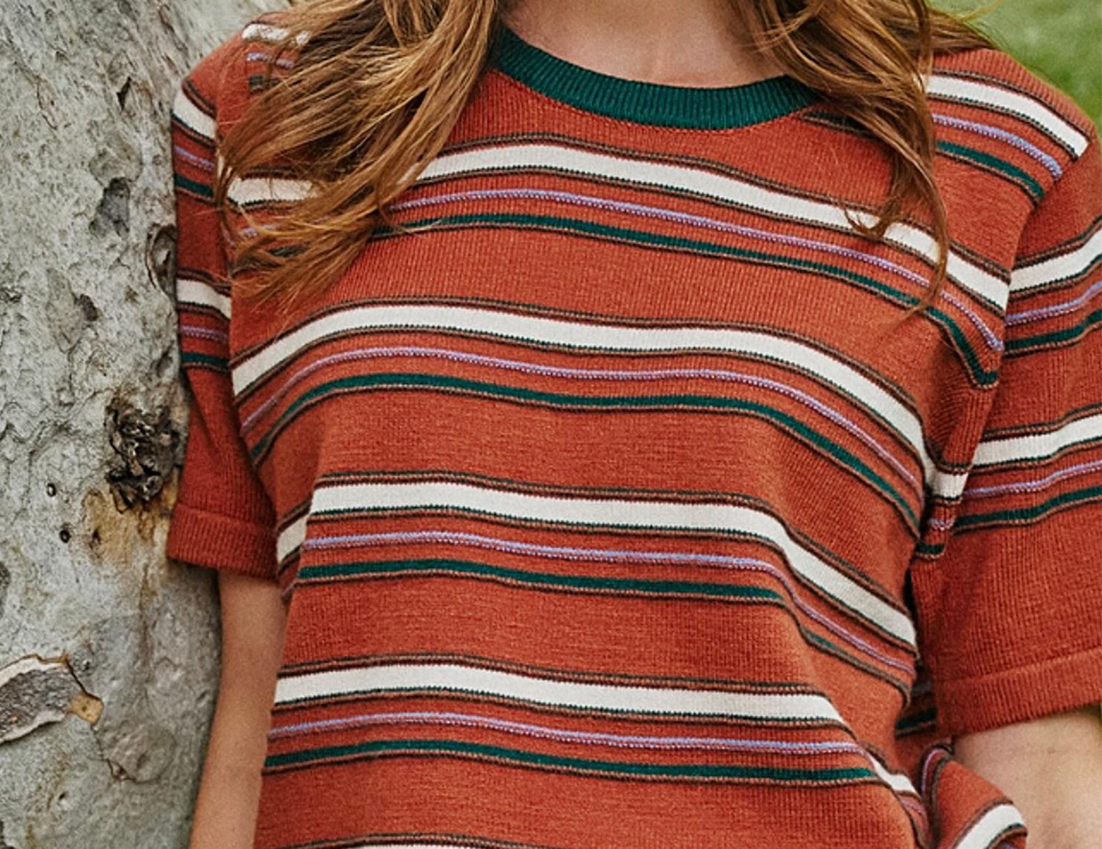 A woman wearing a short-sleeve knit top with horizontal rust orange, white, and dark green stripes, leaning against a textured tree trunk outdoors.