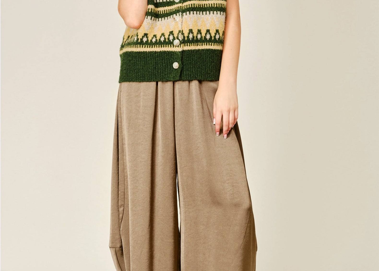 A model wearing a green and beige patterned knit vest with buttons, paired with loose brown wide-leg satin bloomer pants and brown suede mules, against a light beige studio background.